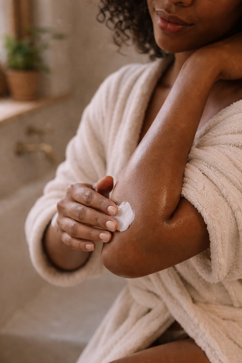 African American woman applying body butter to elbow, smooth glowing skin after using moisturizing body butter for dry skin in warm bathroom setting