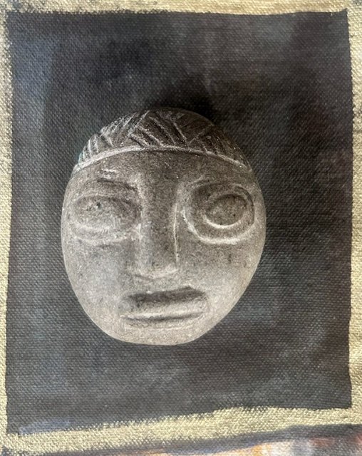 Photograph of an ancient carved stone face mask with large eyes, a prominent nose, and pursed lips on a dark background