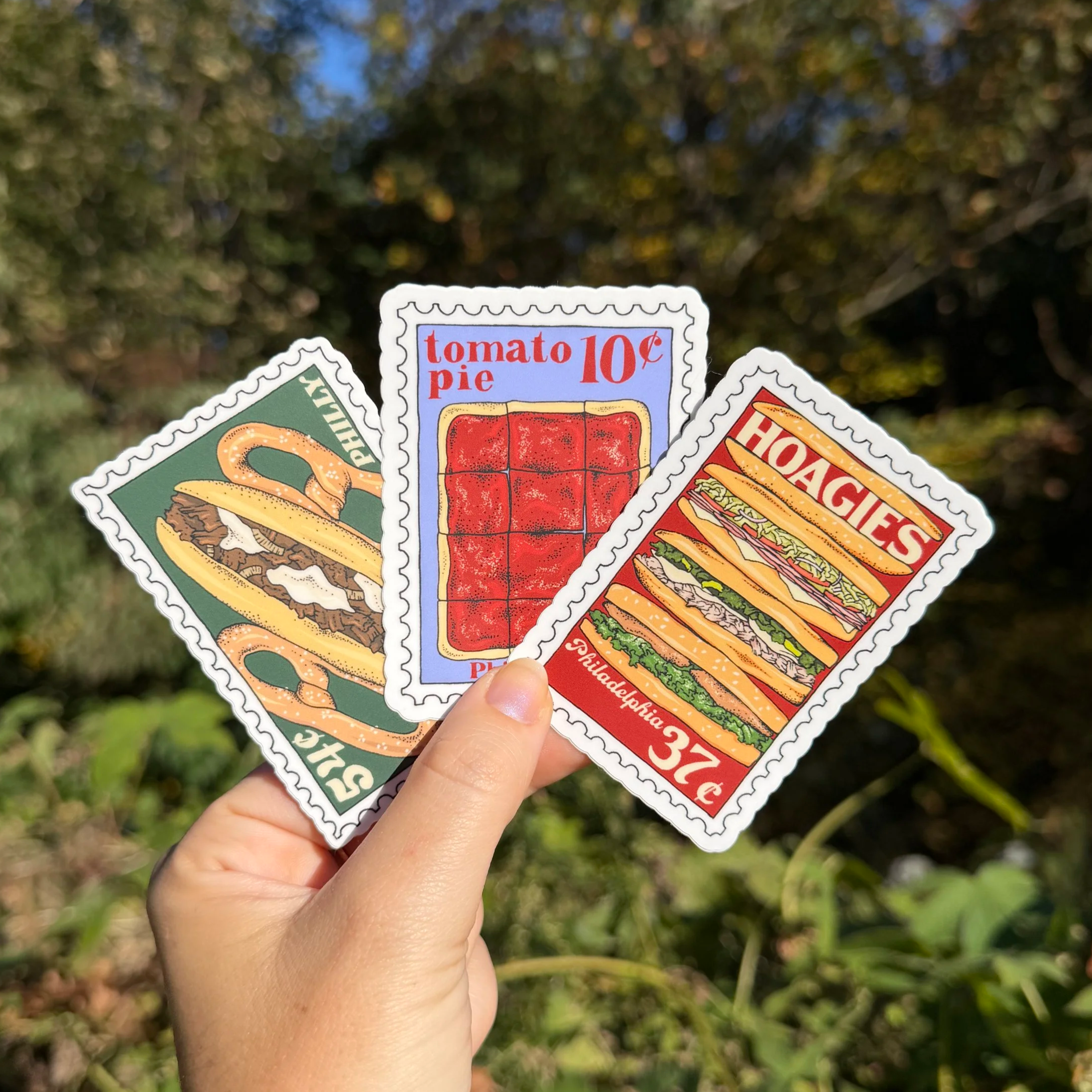Three illustrated stamp stickers. One featuring stack of hoagies, one featuring tomato pie, and one featuring a cheesesteak and soft pretzels.