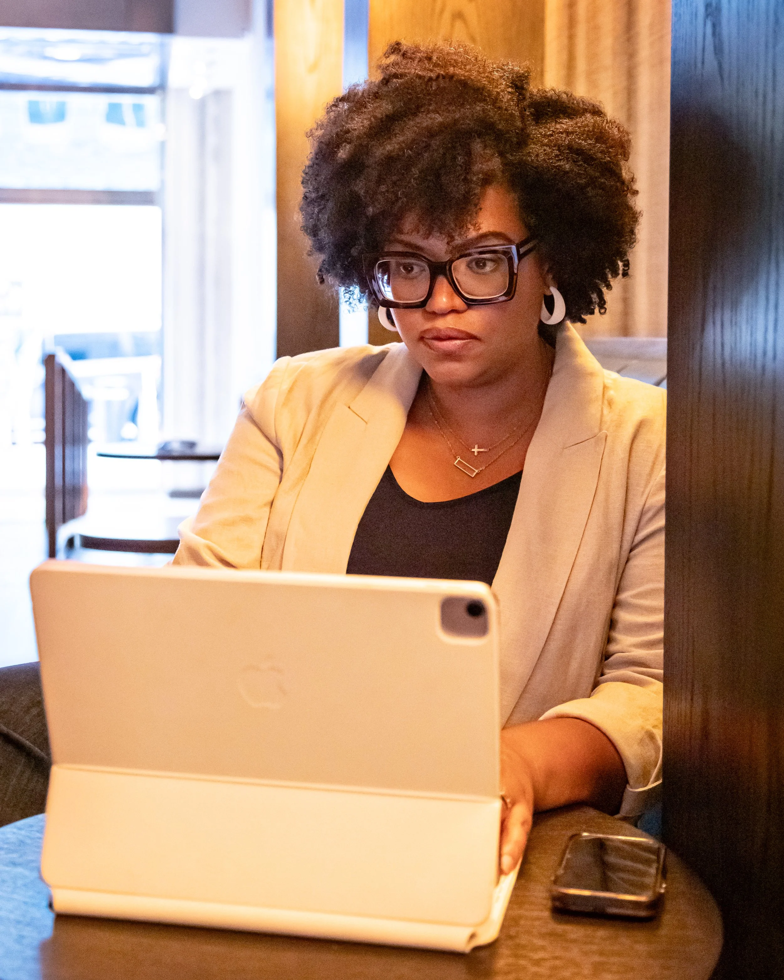 A woman with curly natural hair, glasses, and earrings working on a laptop at a table in a cozy indoor setting, possibly a cafe or restaurant.