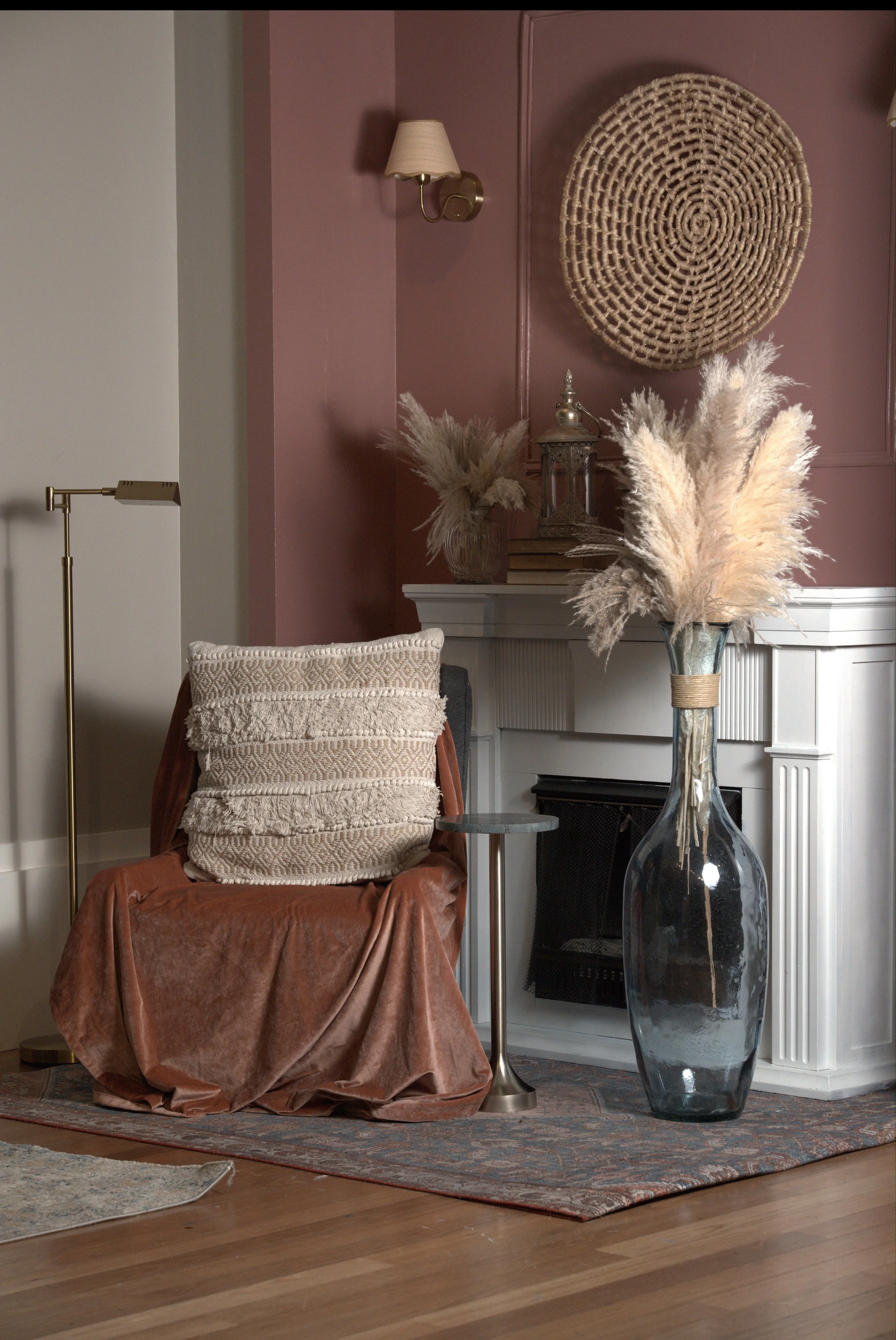 sitting corner with mauve velvet fabric draped over accent chair with a pharmacy lamp on the side and grey stone accent table in front of a white fireplace and mauve painted wall with woven straw basket wall decor