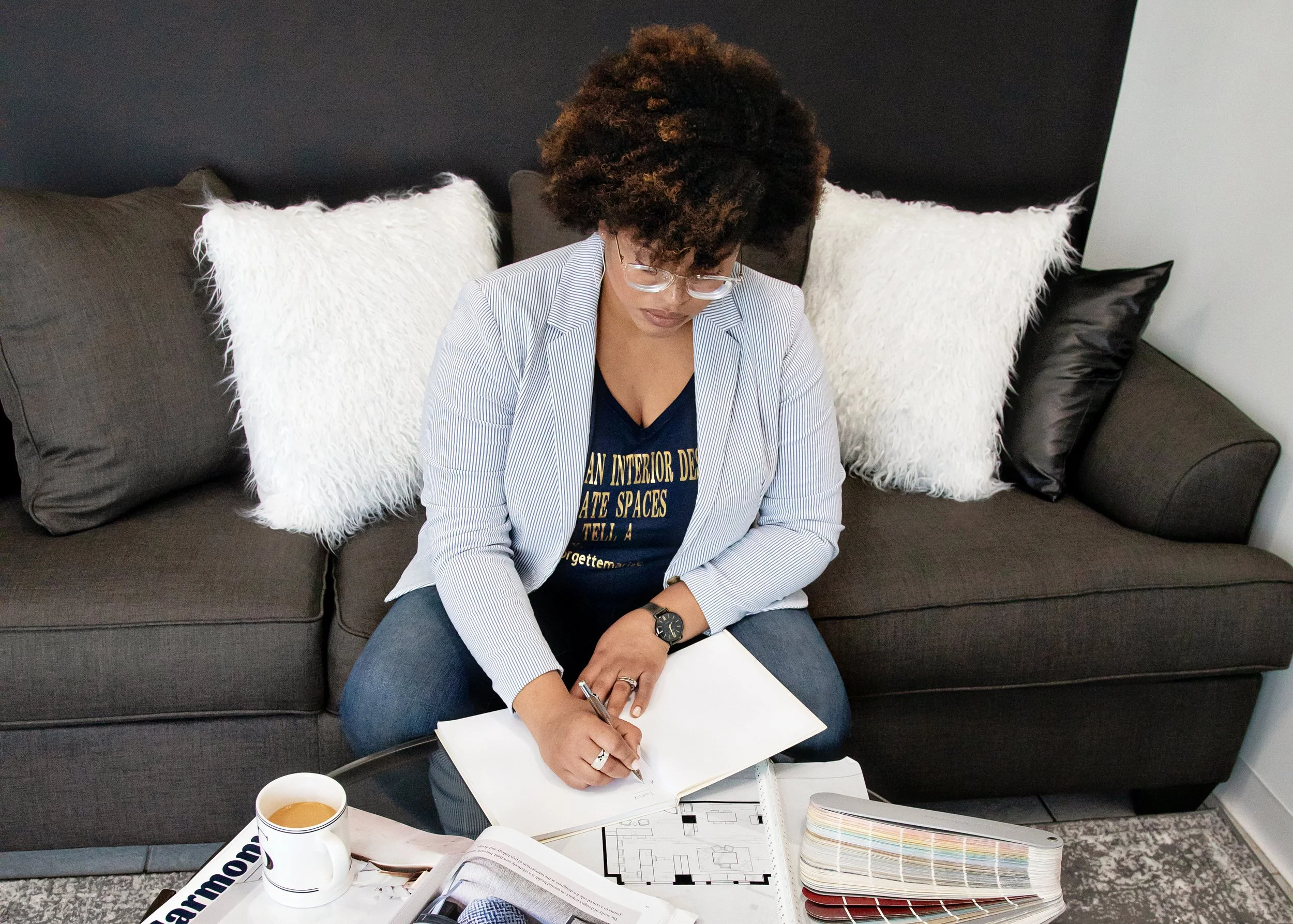 An interior designer sitting on a dark gray couch, writing in a notebook on a small table. She has curly hair, is wearing glasses, a blue shirt, and a light striped blazer. A coffee mug and a color swatch book are on the table in front of her.