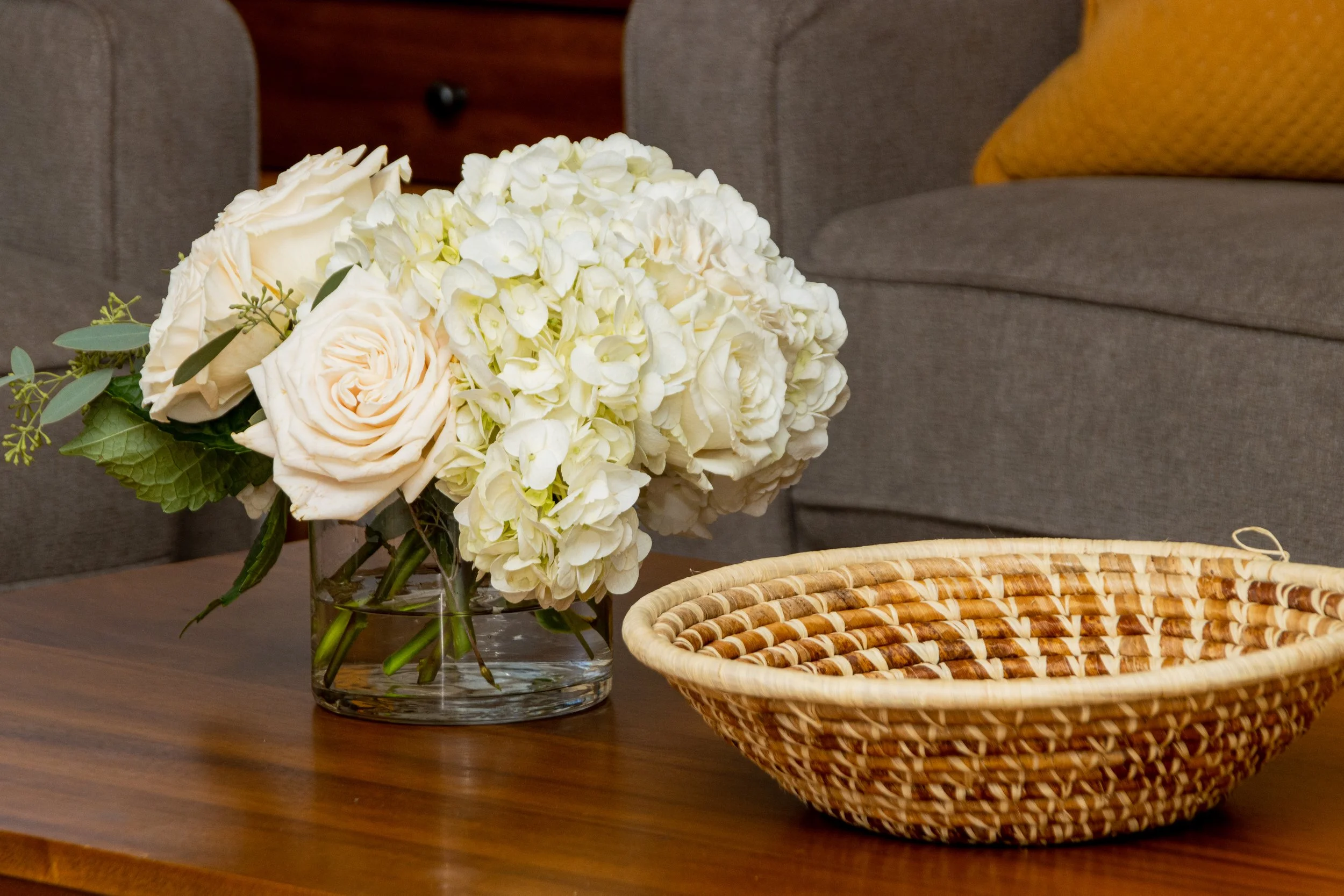 A bouquet of white and cream roses and hydrangeas in a glass vase on a wooden table, next to a woven basket, with a gray couch and yellow pillow in the background.