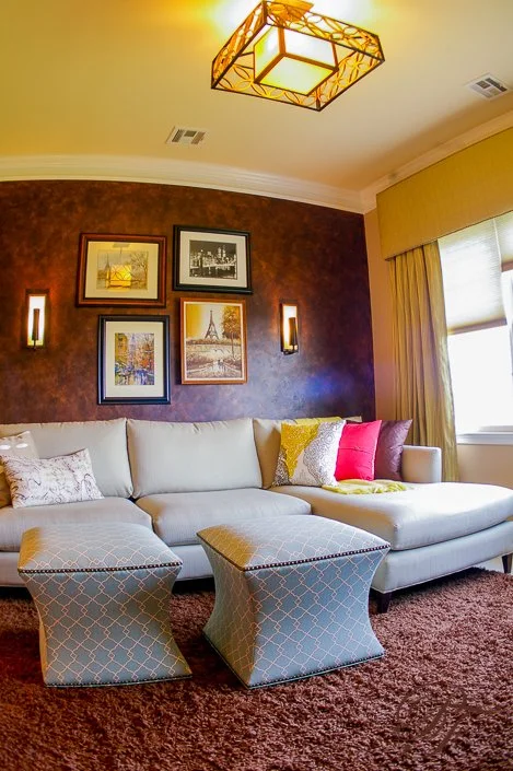 Living room with a beige sectional sofa, two matching ottomans, colorful pillows, artwork on a purple accent wall, a window with curtains, and a decorative ceiling light fixture.