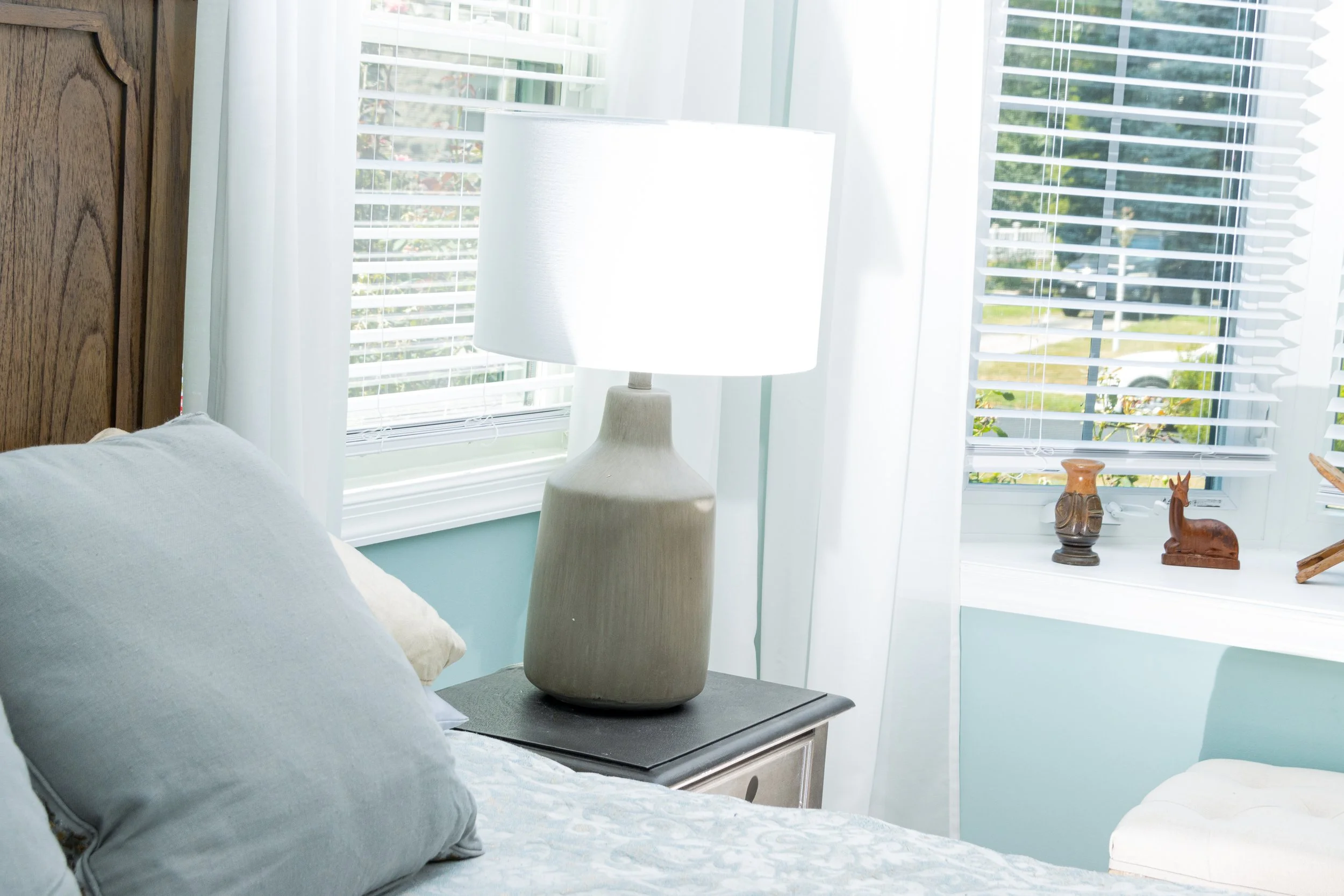 A cozy bedroom corner with a bed, a wooden nightstand, a large beige lamp, and decorative items on the windowsill, with light blue walls and white blinds letting in natural sunlight.