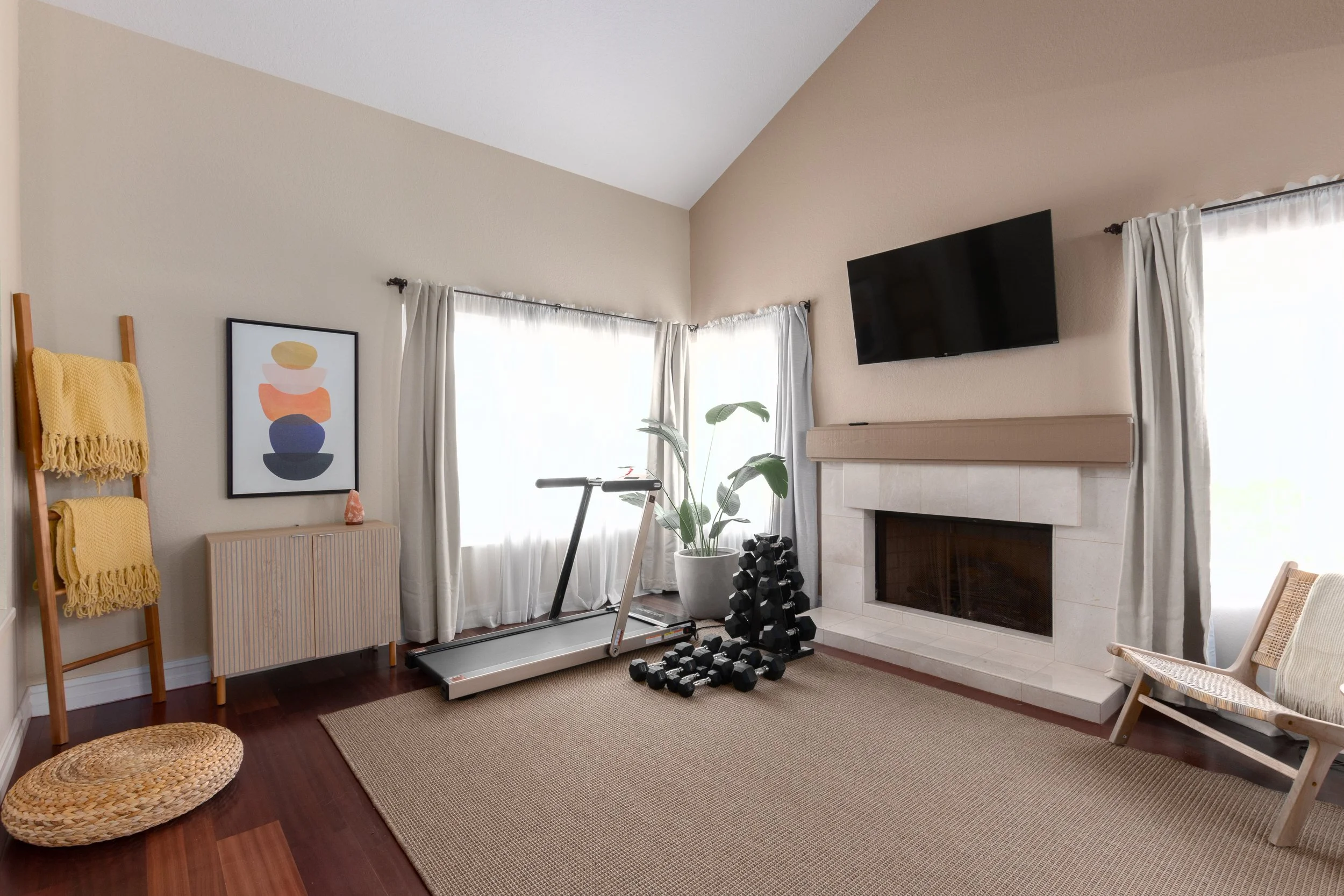 Thoughtfully styled home exercise room combining fitness equipment with soft textures, including a sisal rug, woven cushion, fireplace, and wooden ladder with throws.