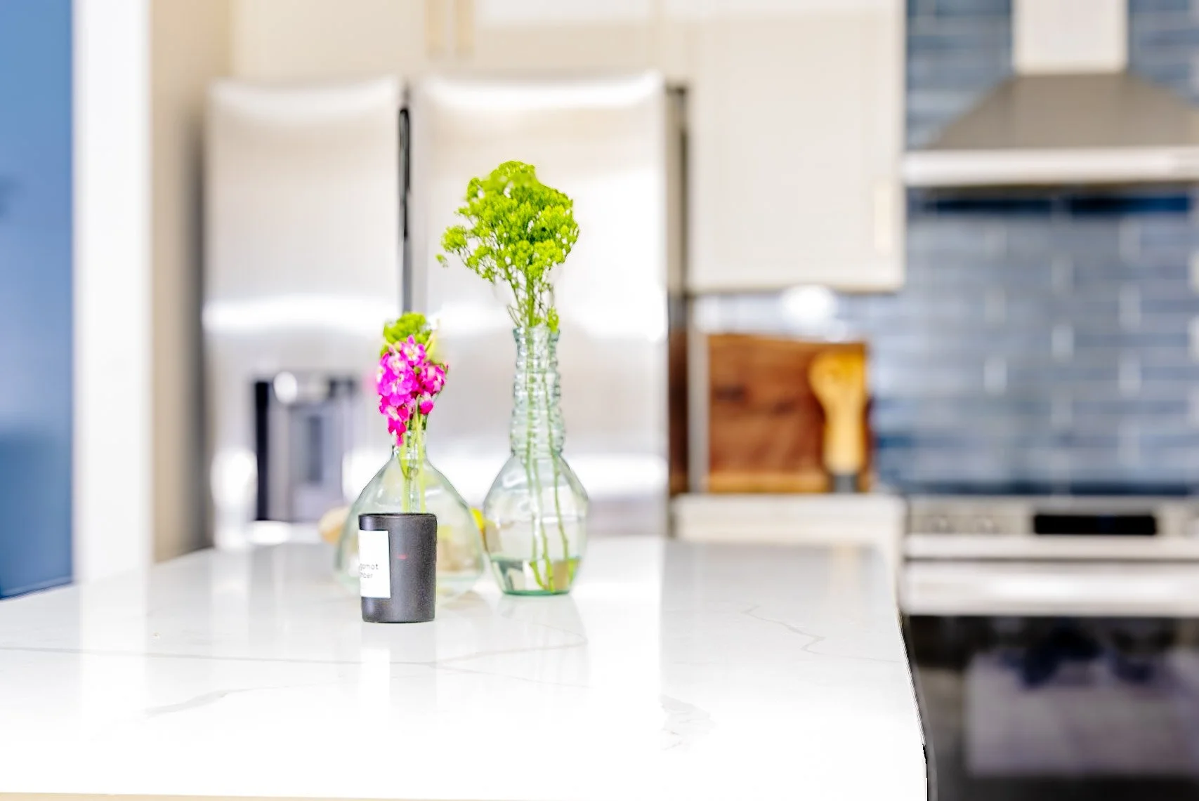 Two glass vases with pink and green flowers on a white kitchen countertop, with a blurred background of kitchen cabinets and appliances.
