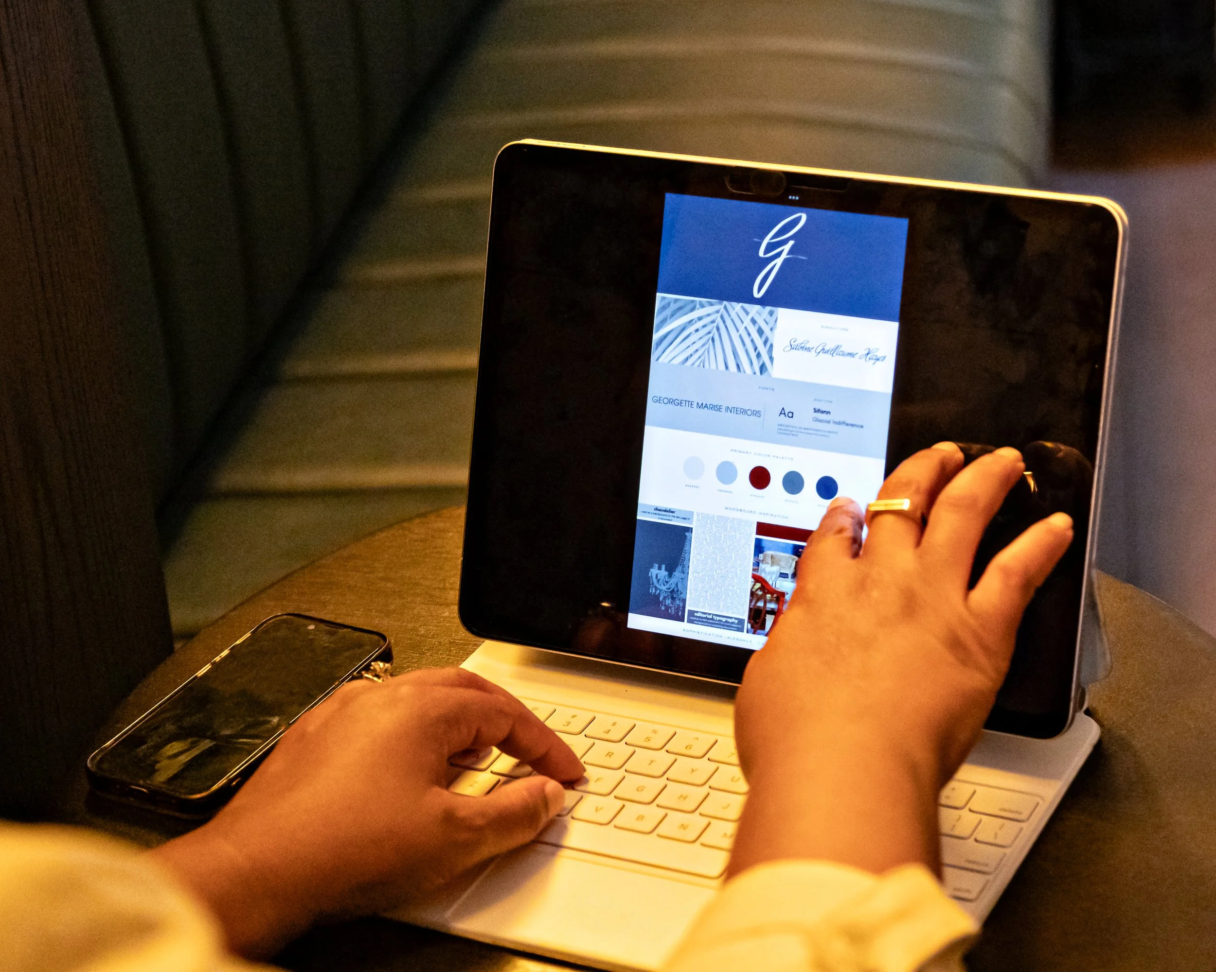 Person using a tablet with a stylus, displaying a design or branding app, with a smartphone on a keyboard case on a table in dim lighting.