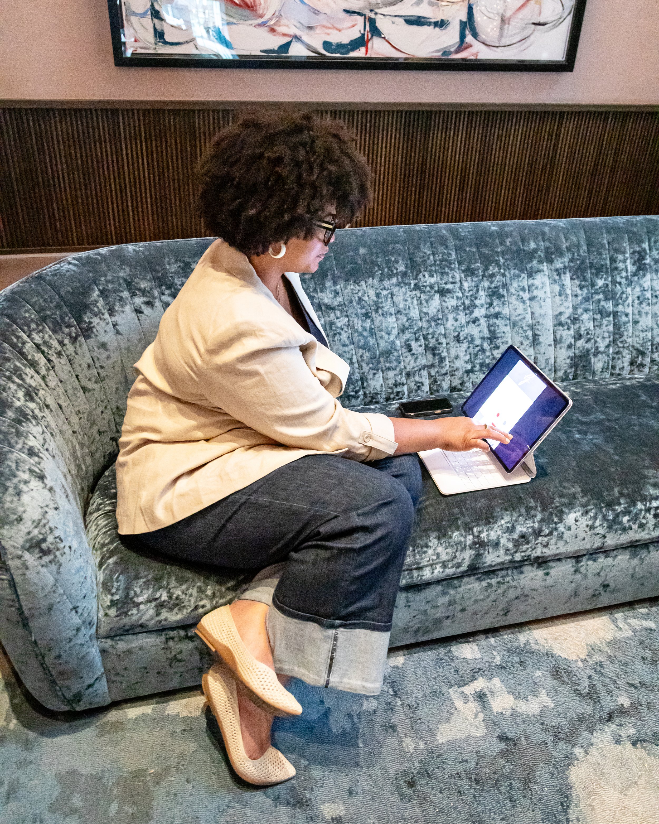 A woman sitting on a vintage sofa, using a tablet computer, with a smartphone next to her on the sofa.