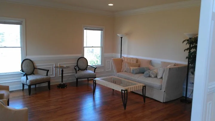 Empty living room with beige walls, hardwood floor, two large windows, a cream-colored sofa with pillows, two upholstered chairs, a coffee table, a floor lamp, and a potted plant.