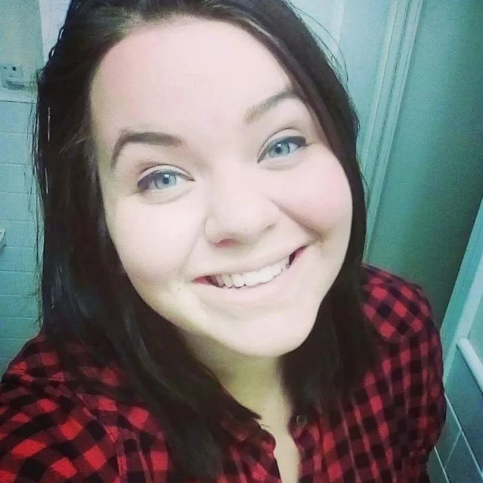 Image Description: A young brunette woman with blue eyes is smiling in a selfie, she is wearing a red plaid flannel and is posed against a green tile wall. 