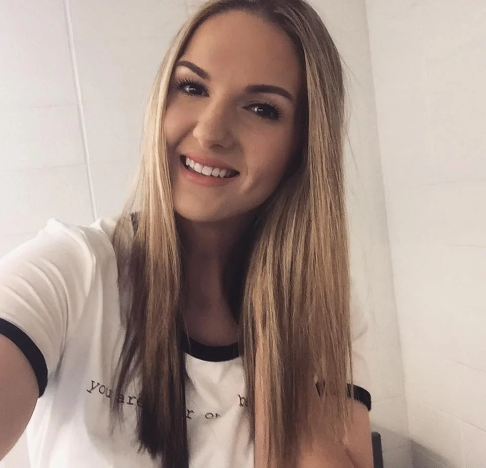 Image Description: A young woman (Carolanne) with long blonde hair, poses in a selfie against a white tiled wall. She is smiling and tilting her head to the right. She is wearing a white t-shirt with unclear lettering. 