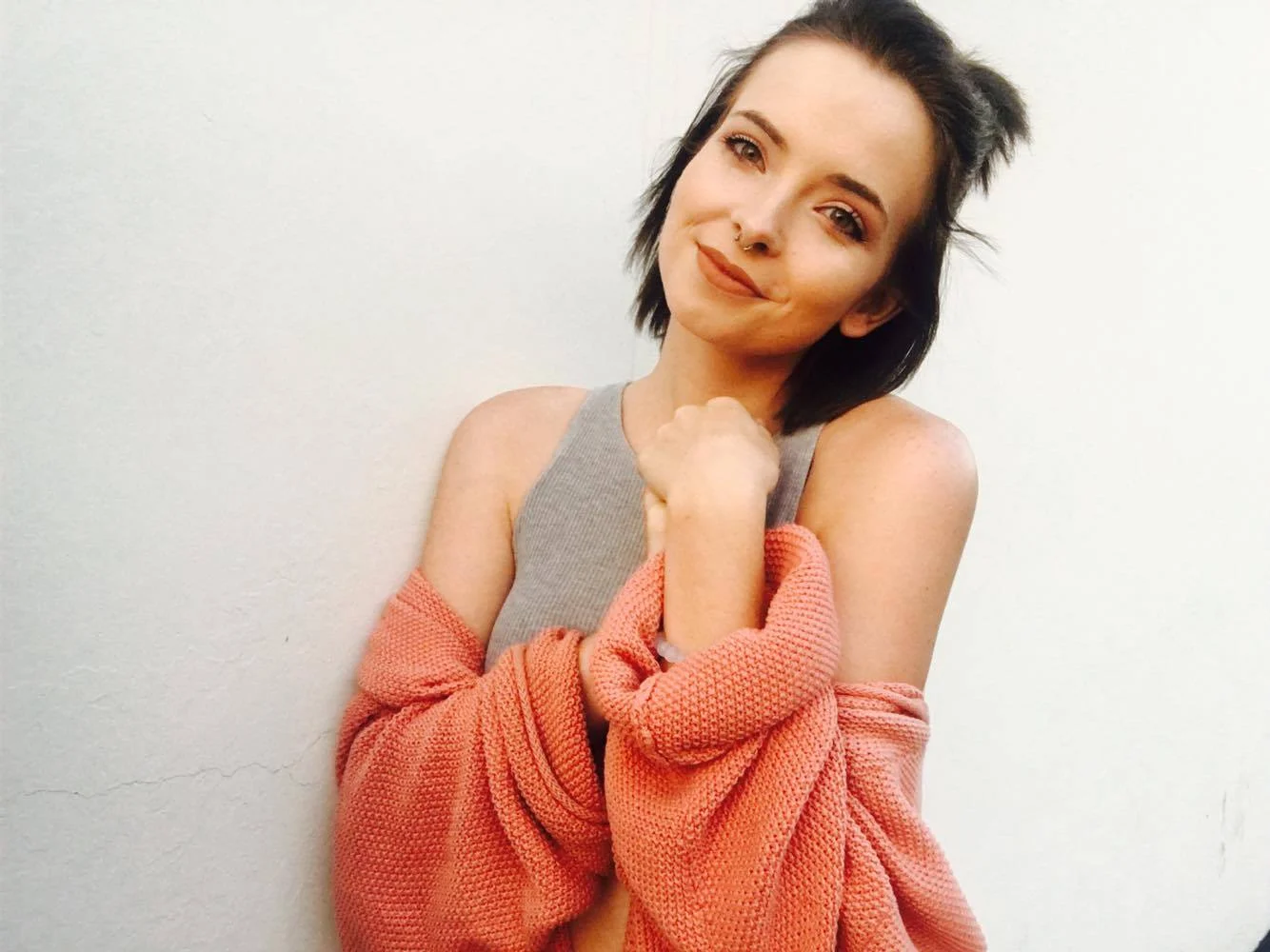 Image Description: A young woman, Ciara, with short brown hair and a septum piercing is smiling in front of a white wall wearing a grey tank top with a coral sweater wrapped around her waist. 