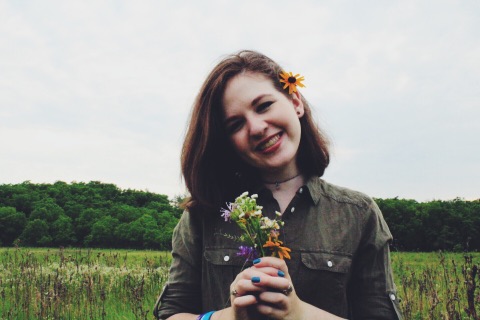 Image Description: A young woman, Katy, smiling with dark brown hair in a field holding a small bouquet of flowers. 