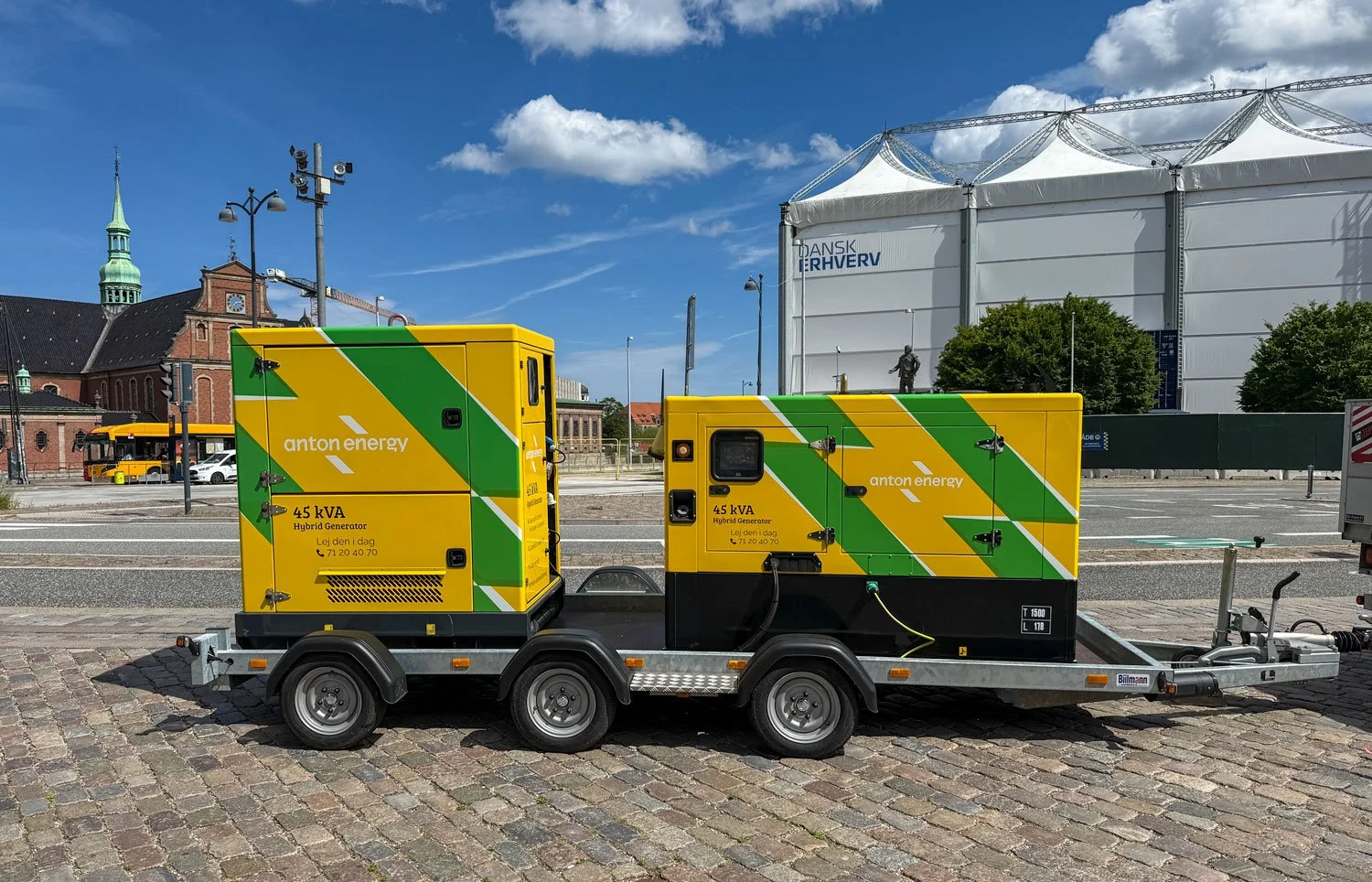 Two yellow and green hybrid generators on a trailer in a city square with historic buildings in the background.