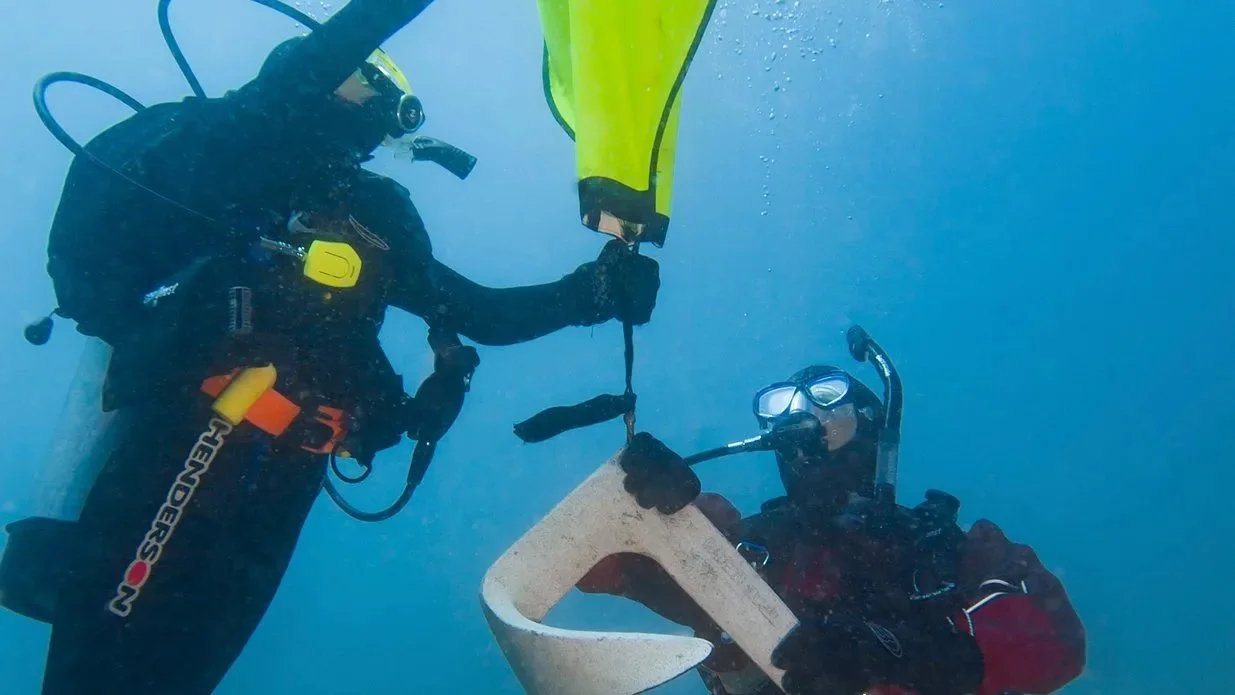Dos buceadores bajo el agua sosteniendo una boya de señalización de superficie amarilla de forma trapezoidal.