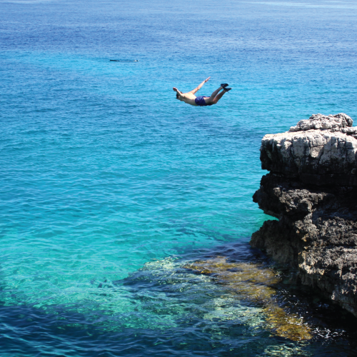 A man dives off of a cliff into the beautiful, clear water below.
