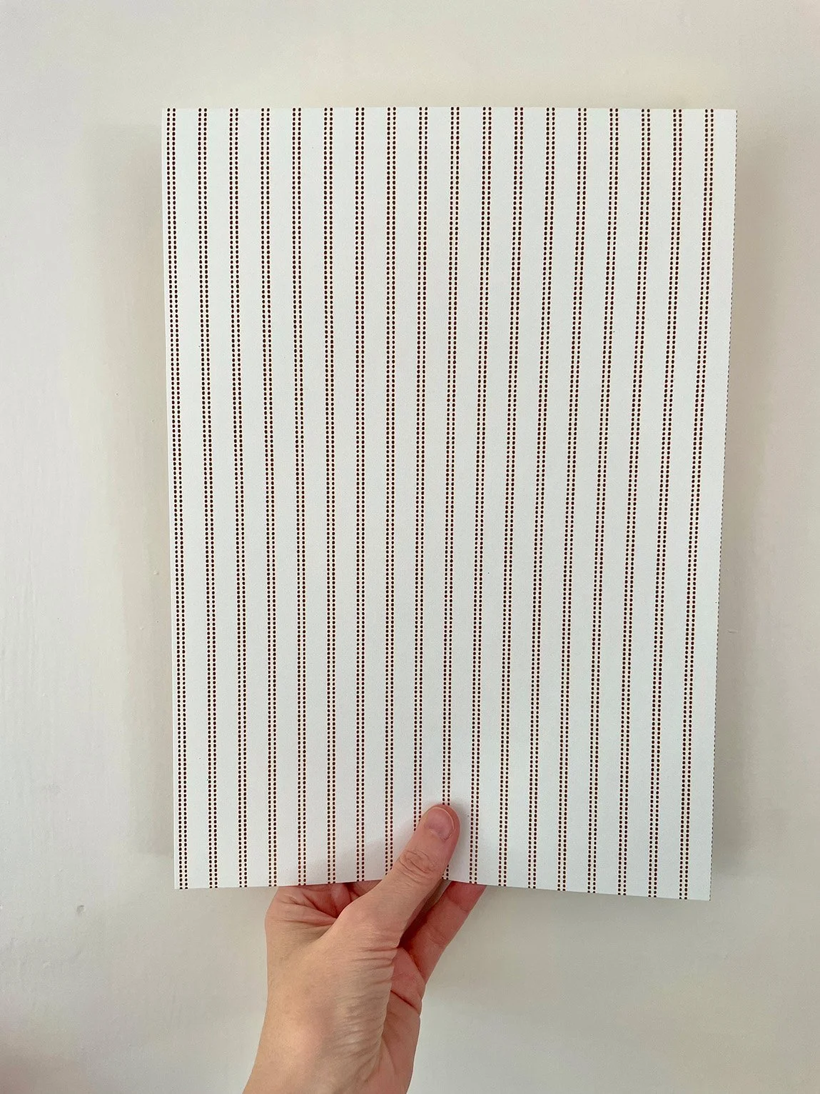 A4 sample of Pillow Stripe wallpaper in Saddle Brown on cream, held against a white wall