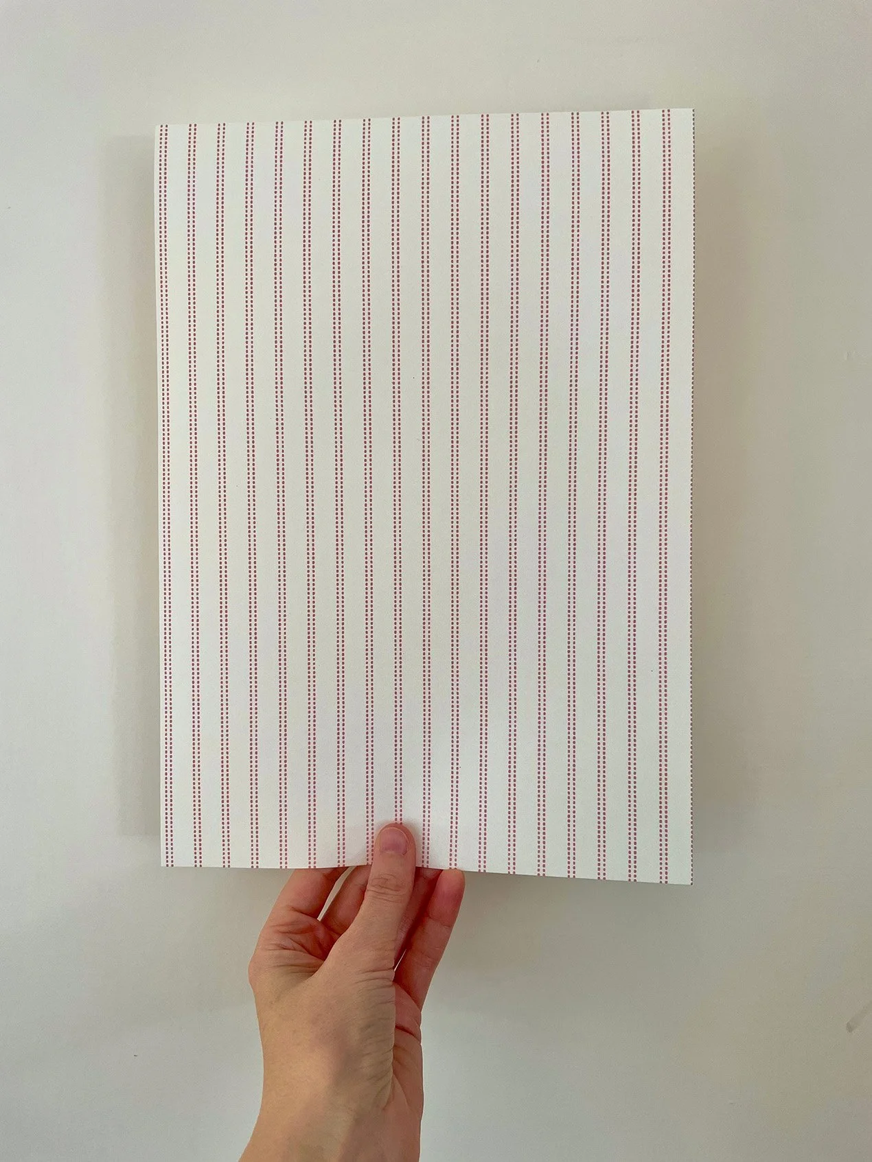 A4 sample of Pillow Stripe wallpaper in Marsala Pink on cream, held against a white wall.