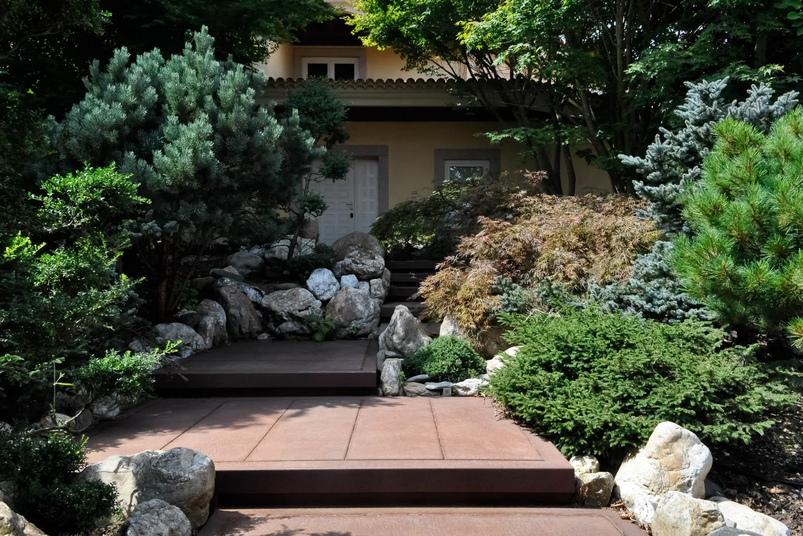 Corten steel walkway steps to a housefront, sided by pines and maples