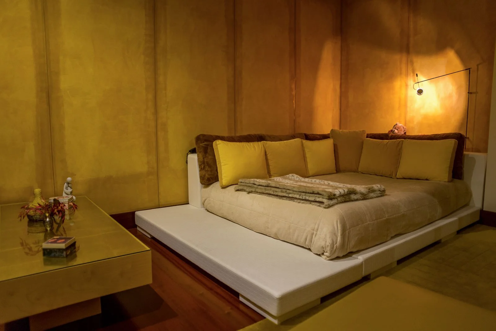 Quarto em tons amarelos com cama, mesa baixa, têxteis e iluminação de autor, e pintura mural decorativa em trompe l'oeil