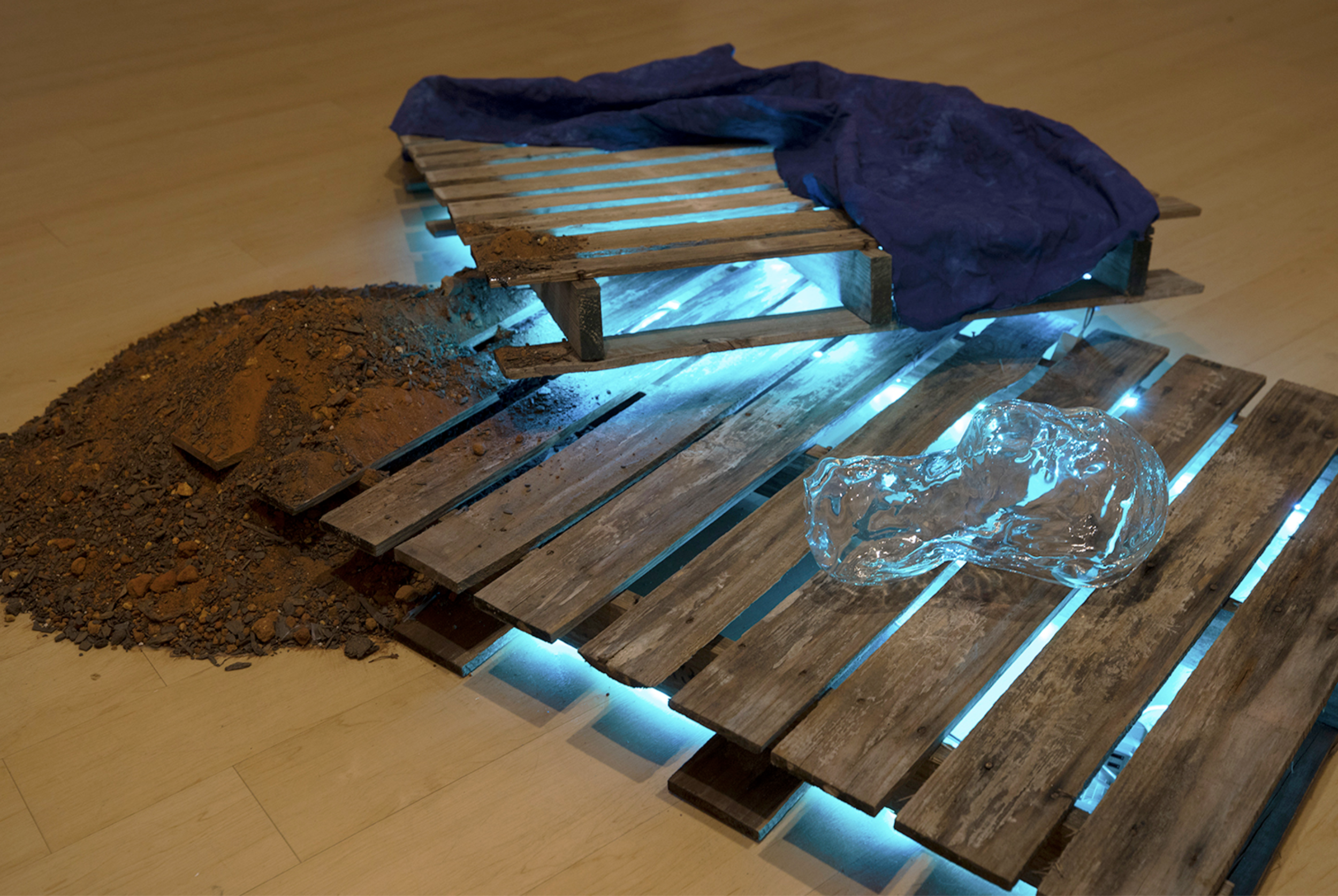  ‘And it all begins in the wake, an after effect’ (2019). Two wooden pallets, blown glass sculpture, oxygen, indigo dye on canvas, red clay dirt, haint-blue led lights 