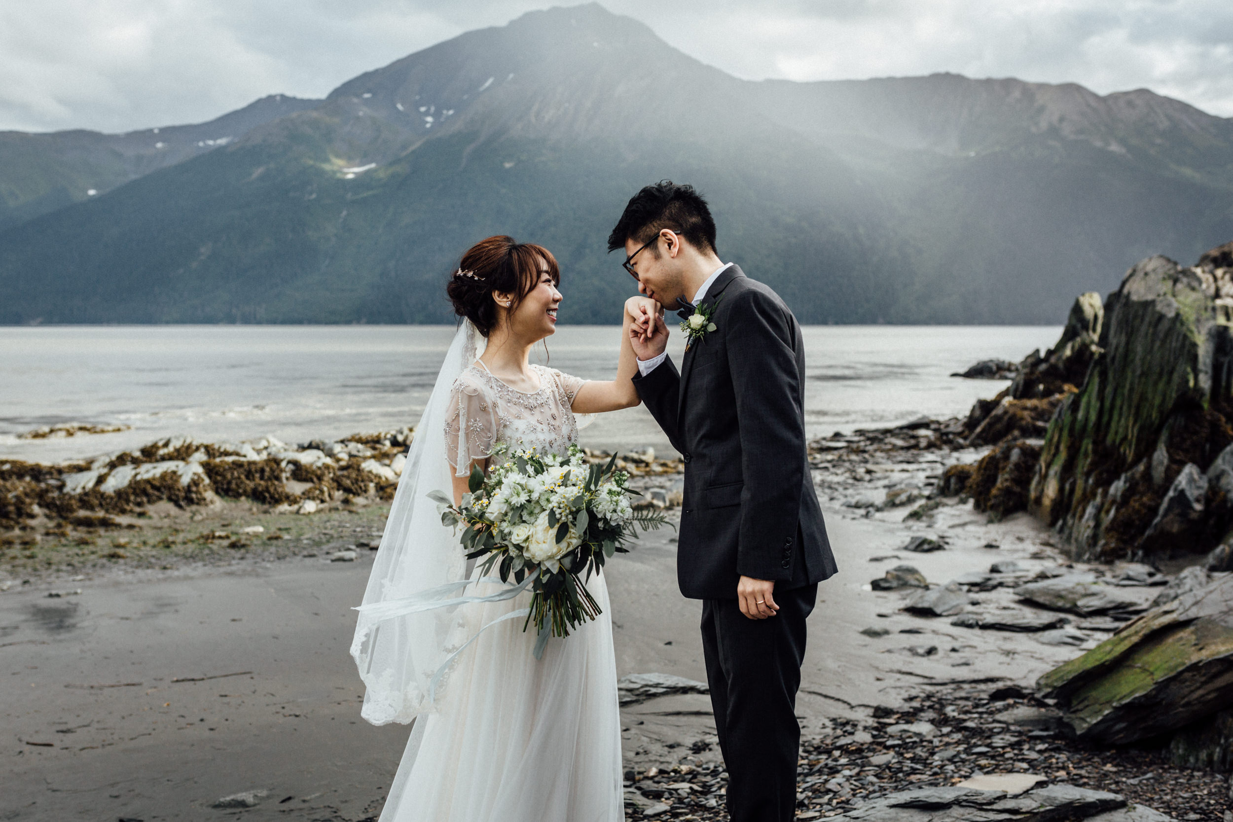 Groom kissing his bride's hand in front of misty mountains