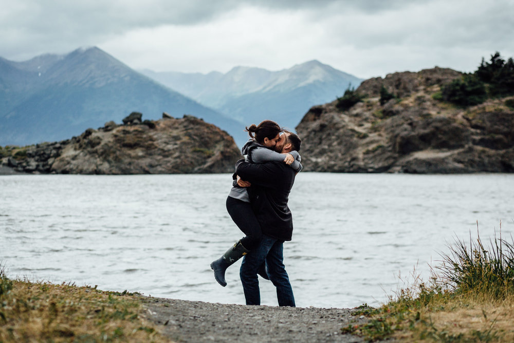 Guy picks up and spins girl in front of water and mountains