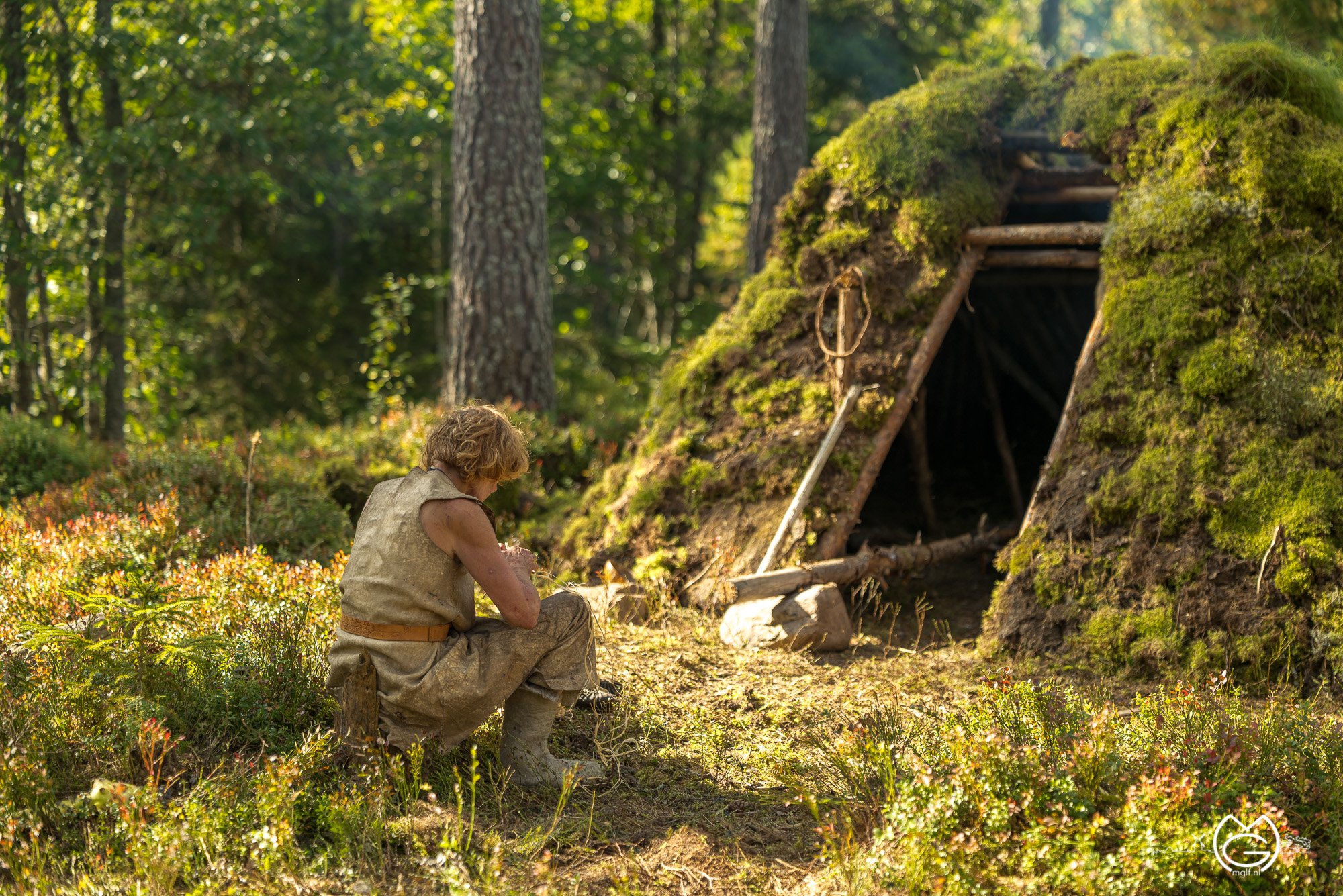 A person sitting next to a moss-covered cave or tent in a dense forest, with trees and greenery surrounding them.