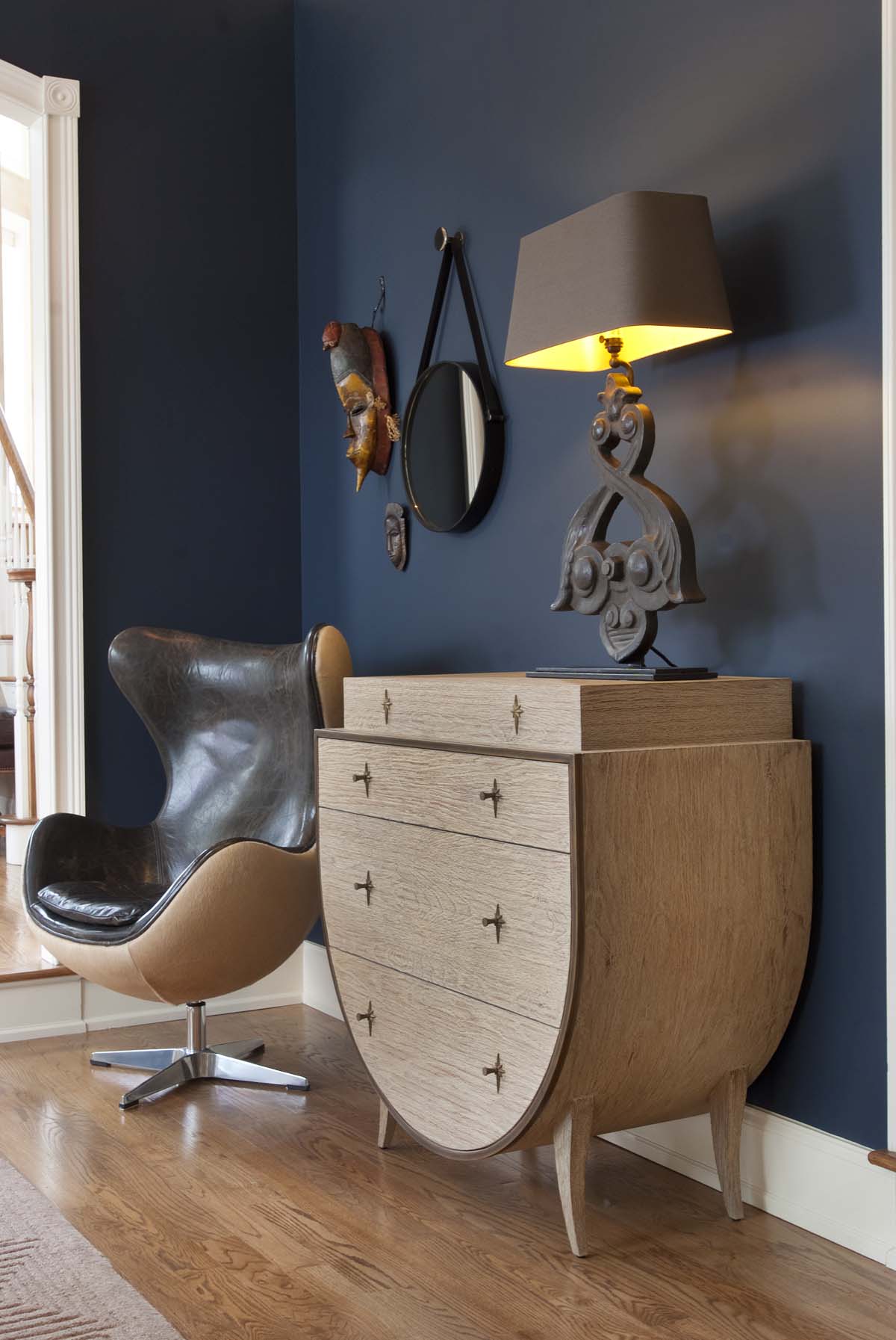 Interior scene with a modern wooden dresser, a leather swivel chair, a table lamp, a circular mirror, a mask, and a decorative mask on a dark blue wall.