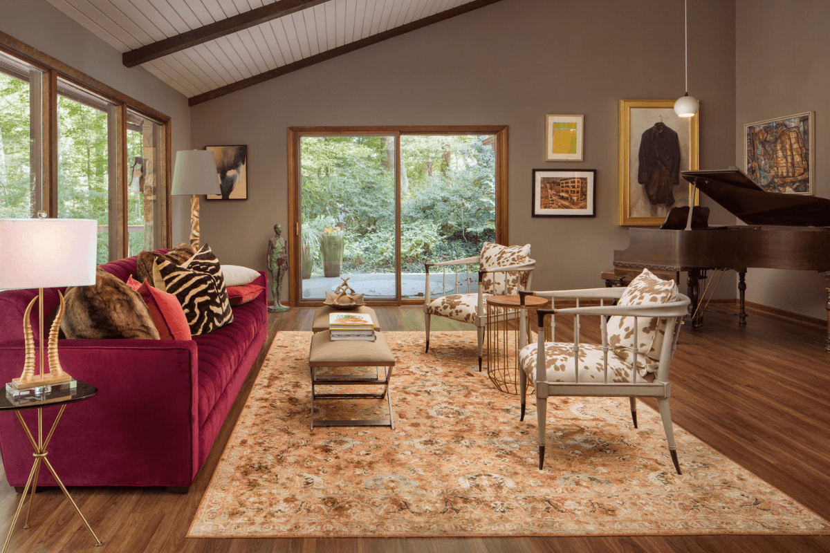 Living room with large windows, gray walls, and a view of greenery outside. Features include a red velvet sofa with animal print pillows, a modern glass and gold side table, a beige ottoman, two white armchairs with beige and white patterned cushions, a patterned area rug, a grand piano, and various artworks on the wall.