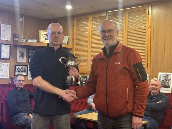 Photo of Chris Stirling the 2025-26 Speedway Winner receiving the Bill Dick Trophy from Robin Thomson in the Club Room at Belerno and Currie Rifle Club