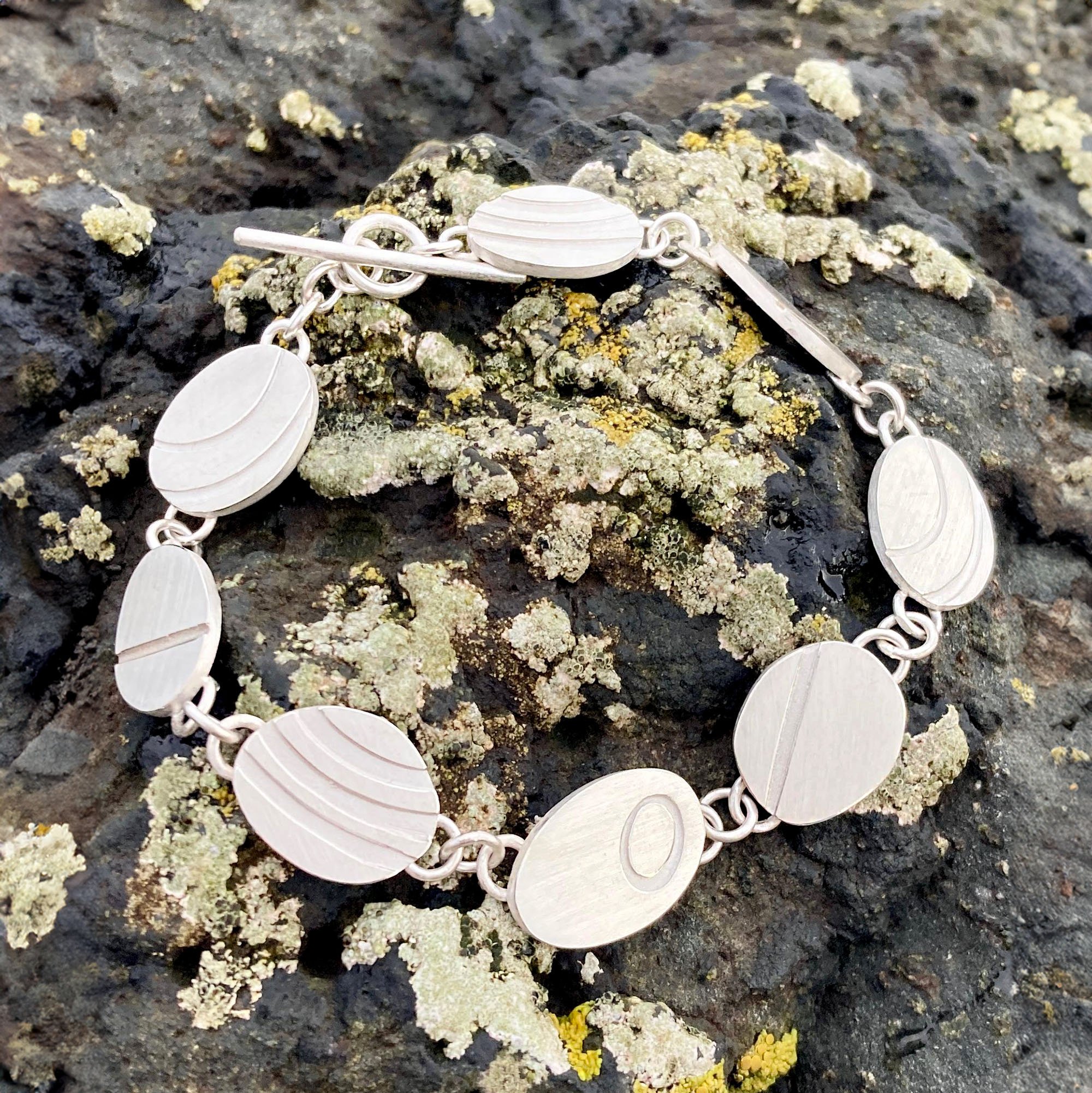 Silver pebble bracelet with tactile charms, inspired by the natural quartz lines of Cornish beach stones, displayed on a rock at the beach.