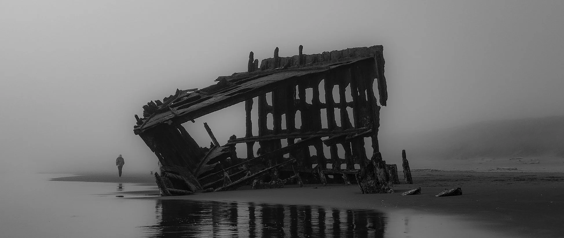 An old burned structure, maybe a ship, on a beach with a lone many walking nearby.