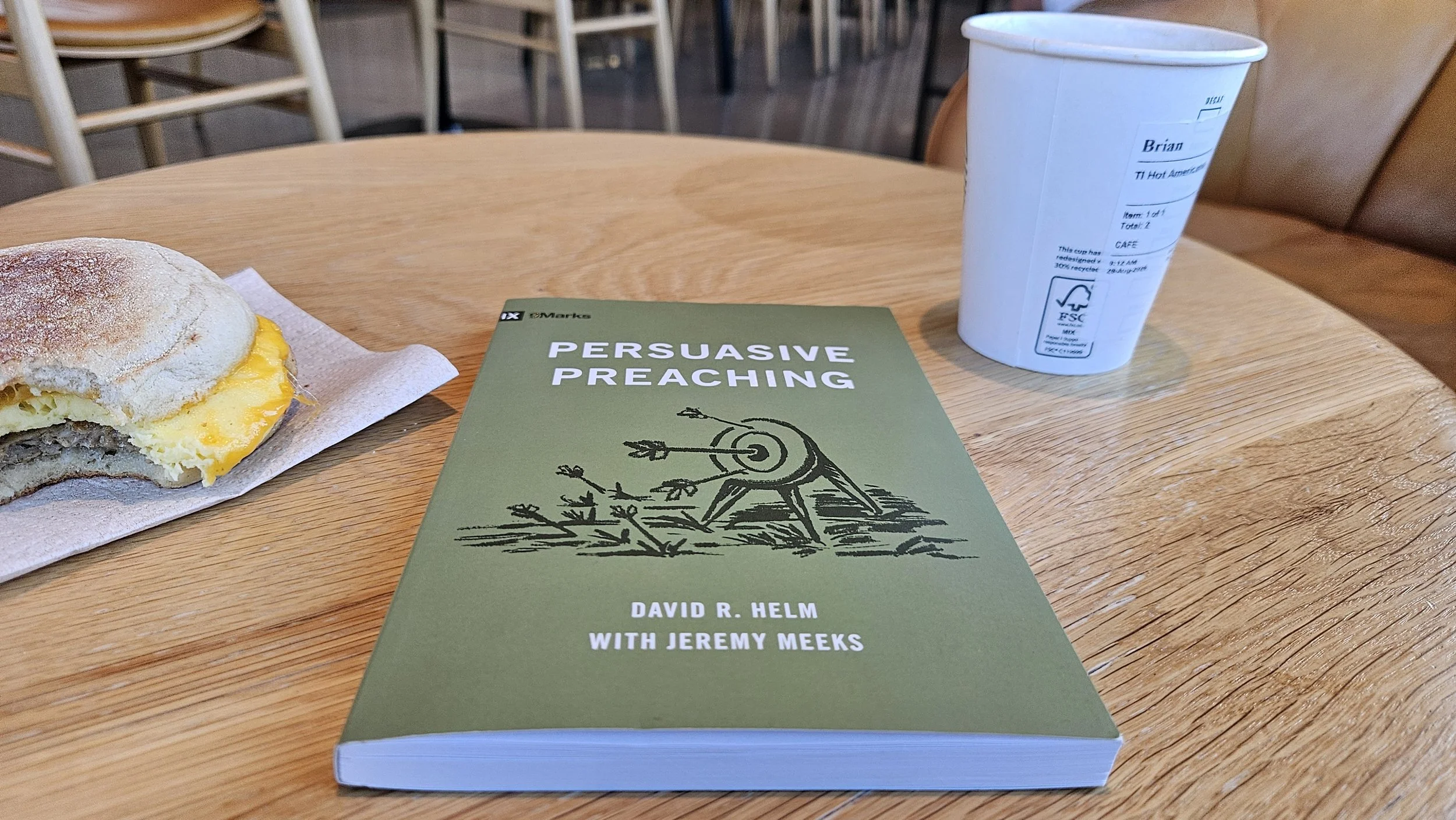 The book, Persuasive Preaching by David Helm on a table with an egg sandwich and cup of coffee.