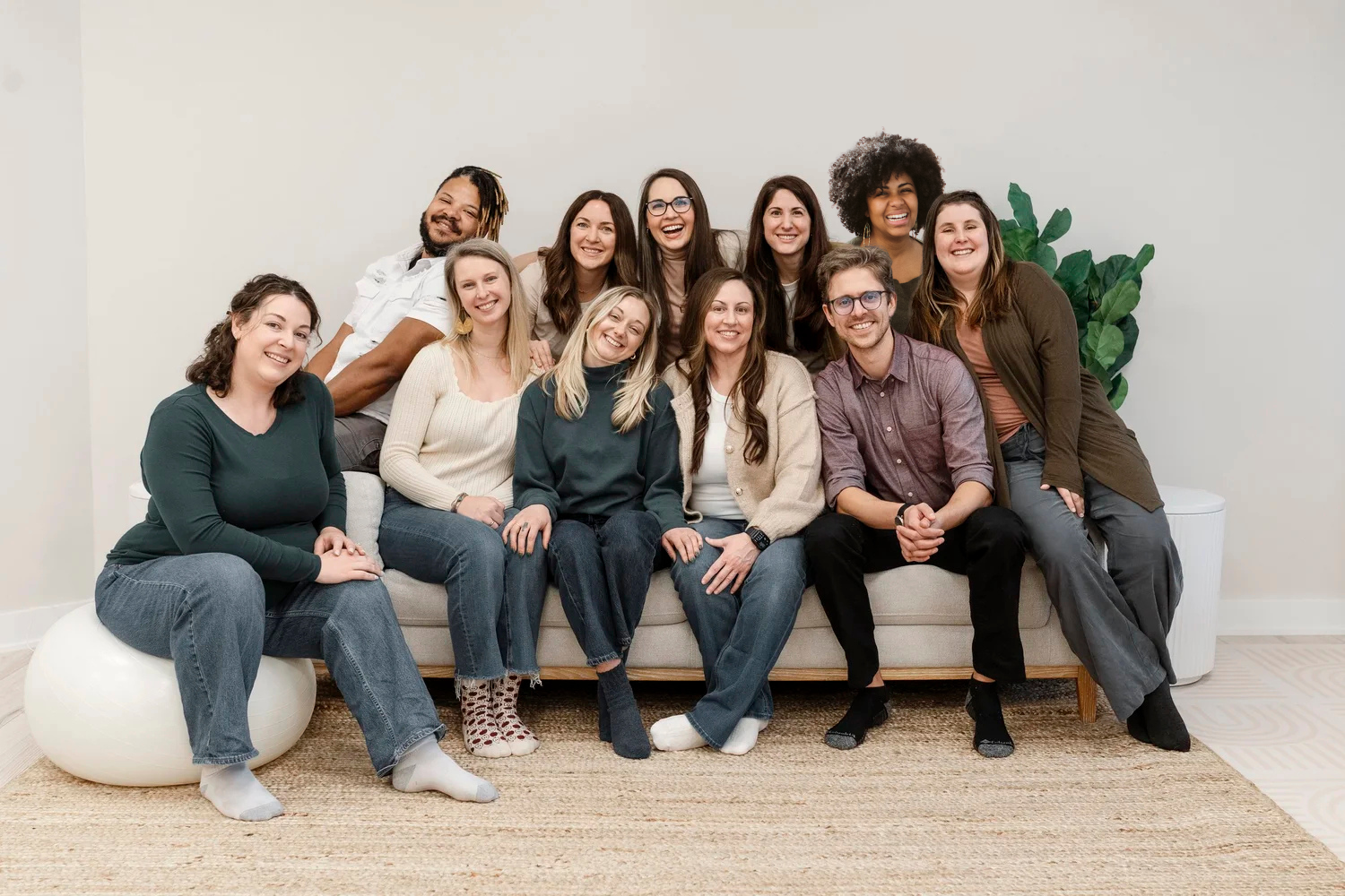 A diverse group of 13 smiling Portland perinatal practicioners gathered together on and around a beige sofa in a bright room with a white wall and green plants in the background.