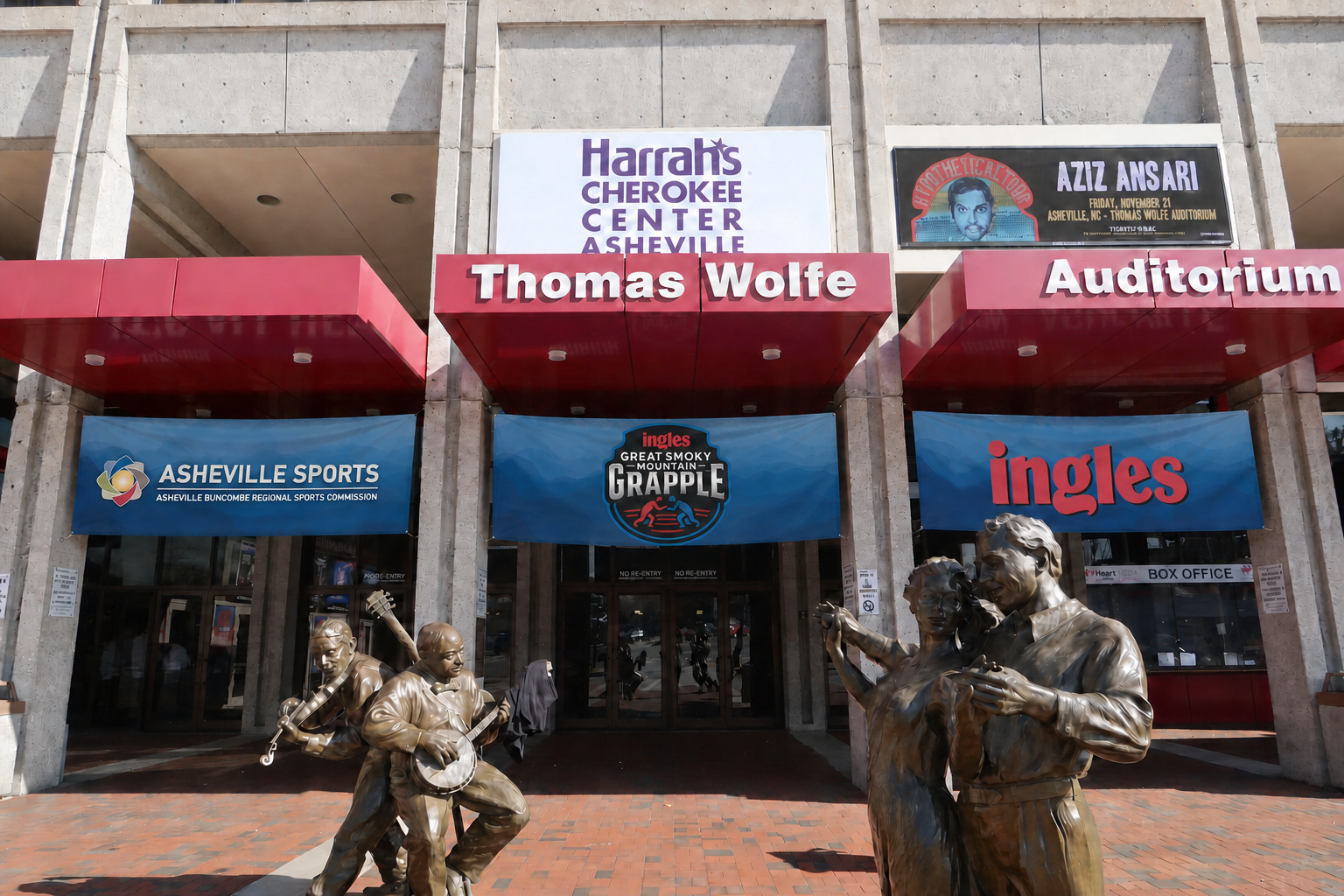 Exterior of the Thomas Wolfe Auditorium at the Harrah's Cherokee Center Asheville, with statue sculptures of musicians in front, banners for sports and entertainment events, and signage for Asheville Sports and Ingles.