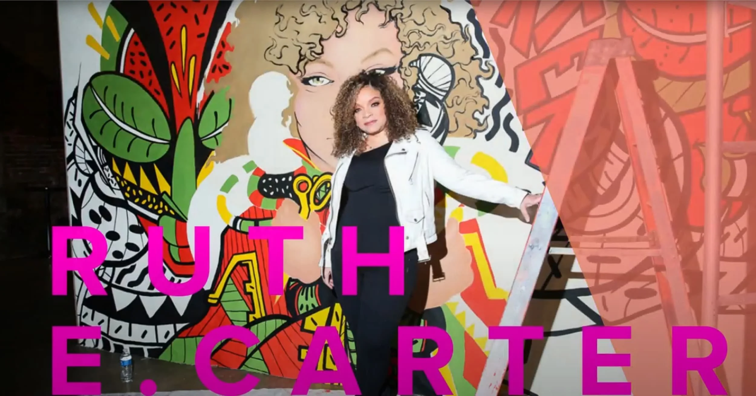 A woman with curly hair standing in front of a colorful graffiti mural, holding a large pink sign that reads 'RUTH E. CARTER'.