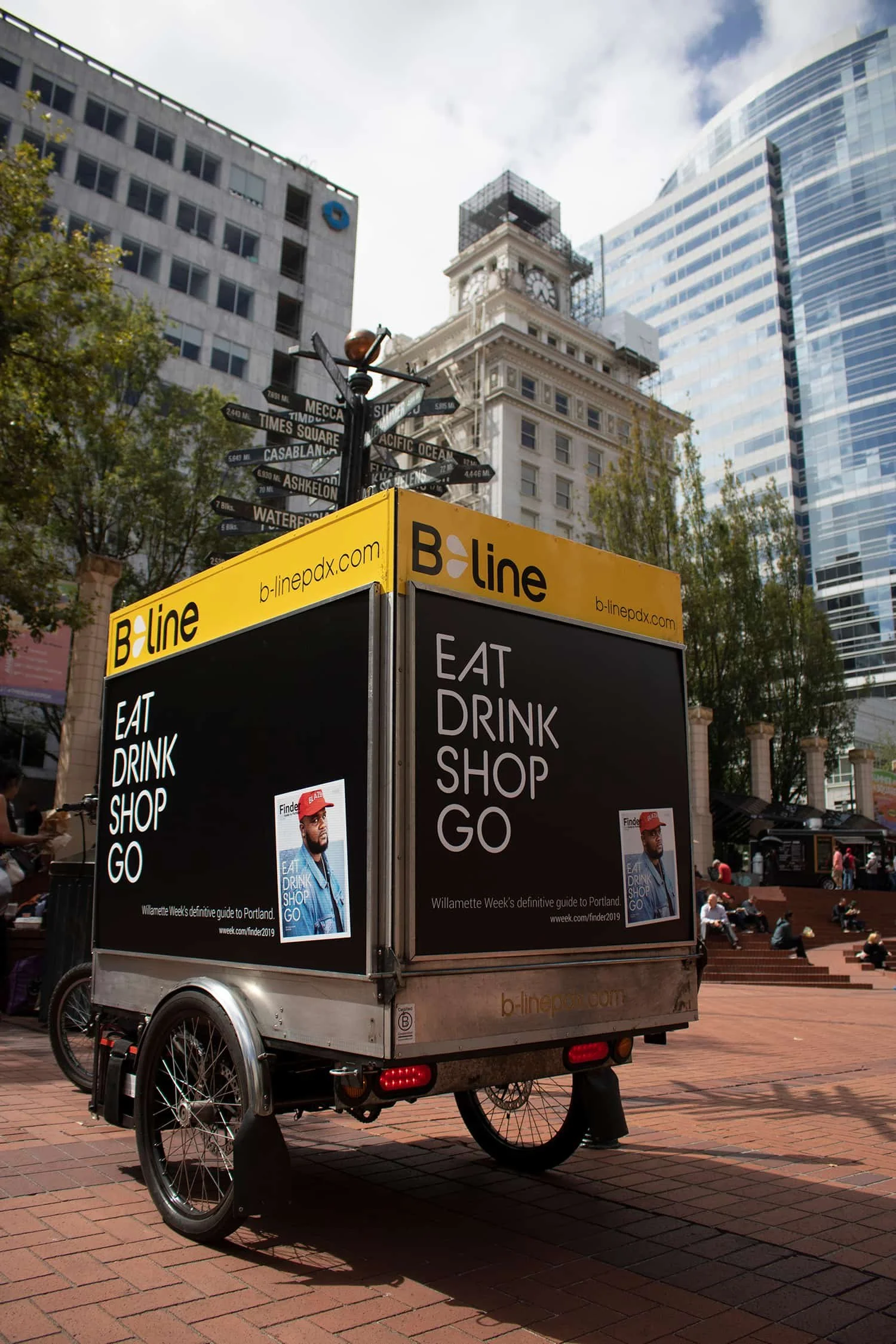 A bike-powered food cart with a yellow sign displaying 'EAT, DRINK, SHOP, GO' in a public outdoor plaza. In the background, tall modern buildings and a historic clock tower can be seen, along with some trees and people sitting on steps.