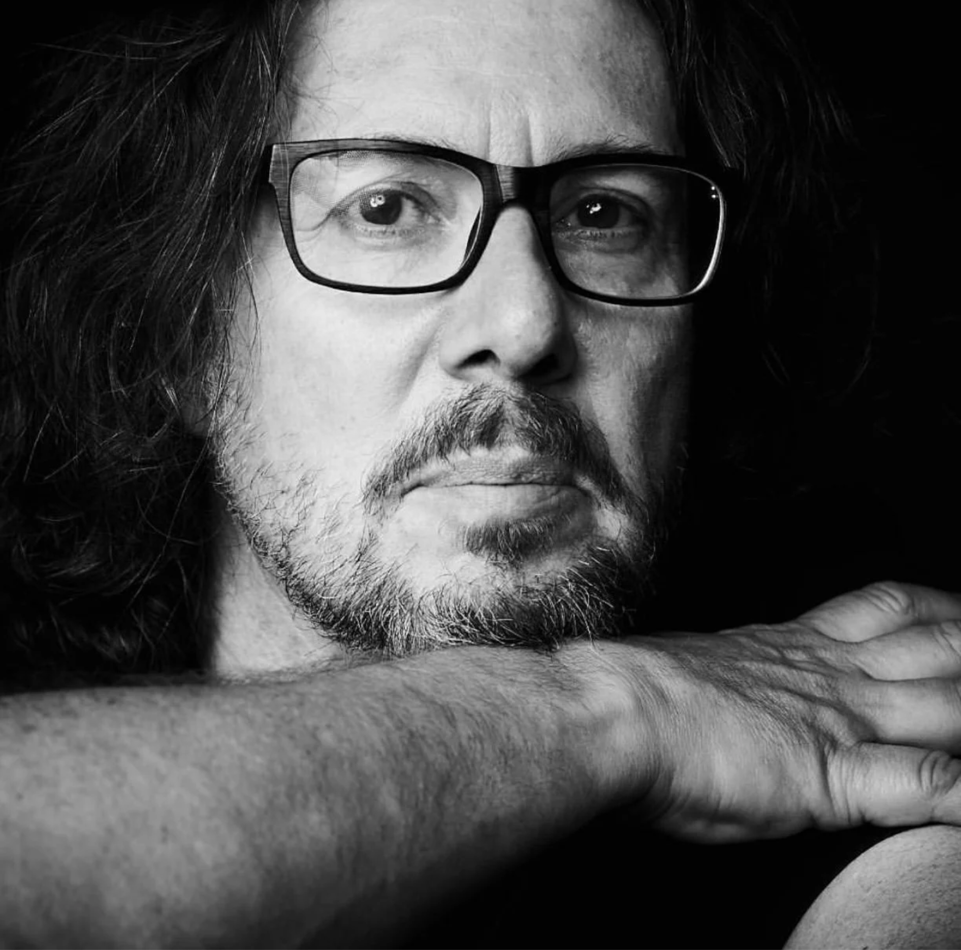 Black and white photo of a person sitting on a patterned rug, wearing a long-sleeve shirt and pants, resting their chin on their hand.