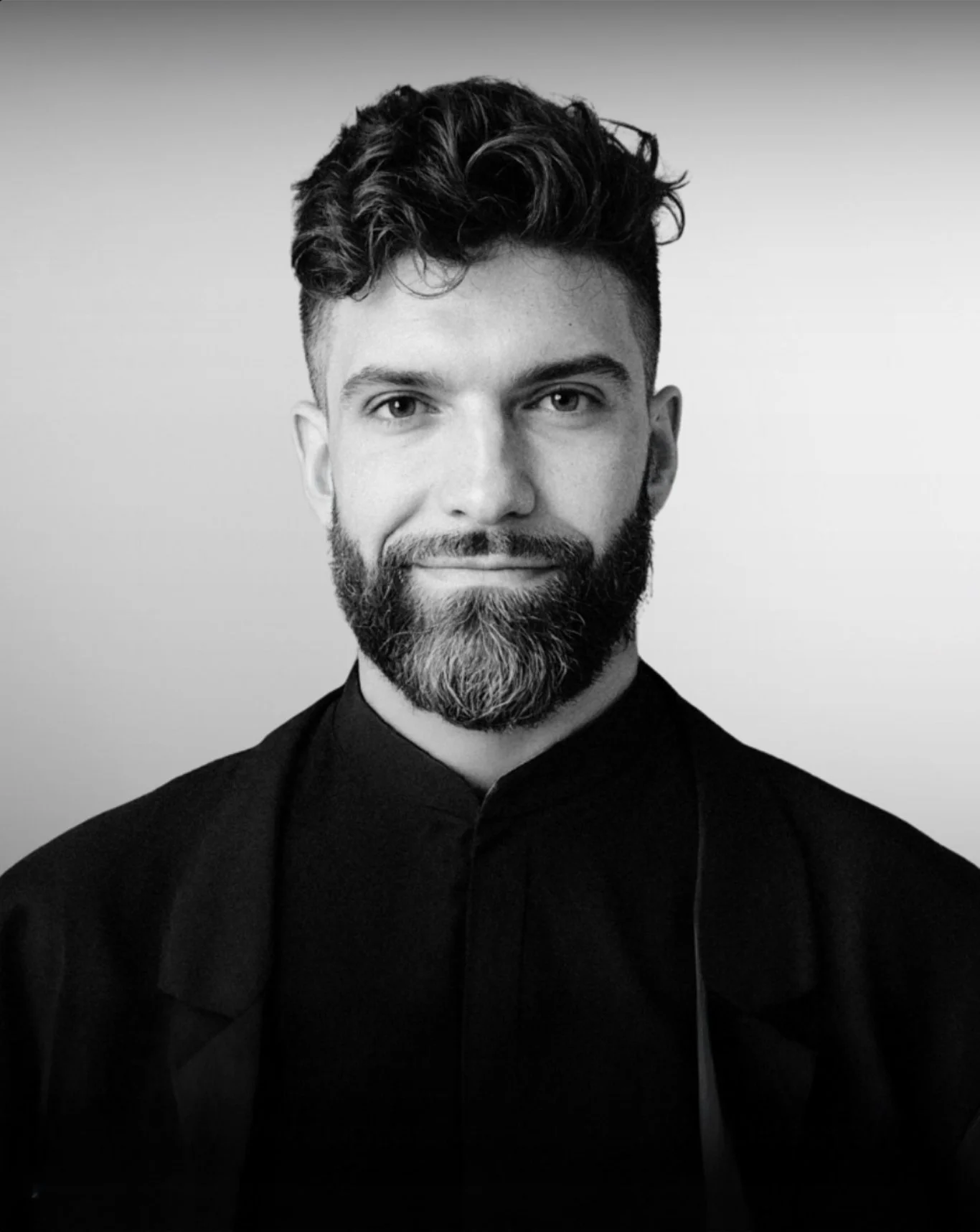 Black and white photo of a person sitting on a cushion on the floor, wearing casual clothes and smiling.