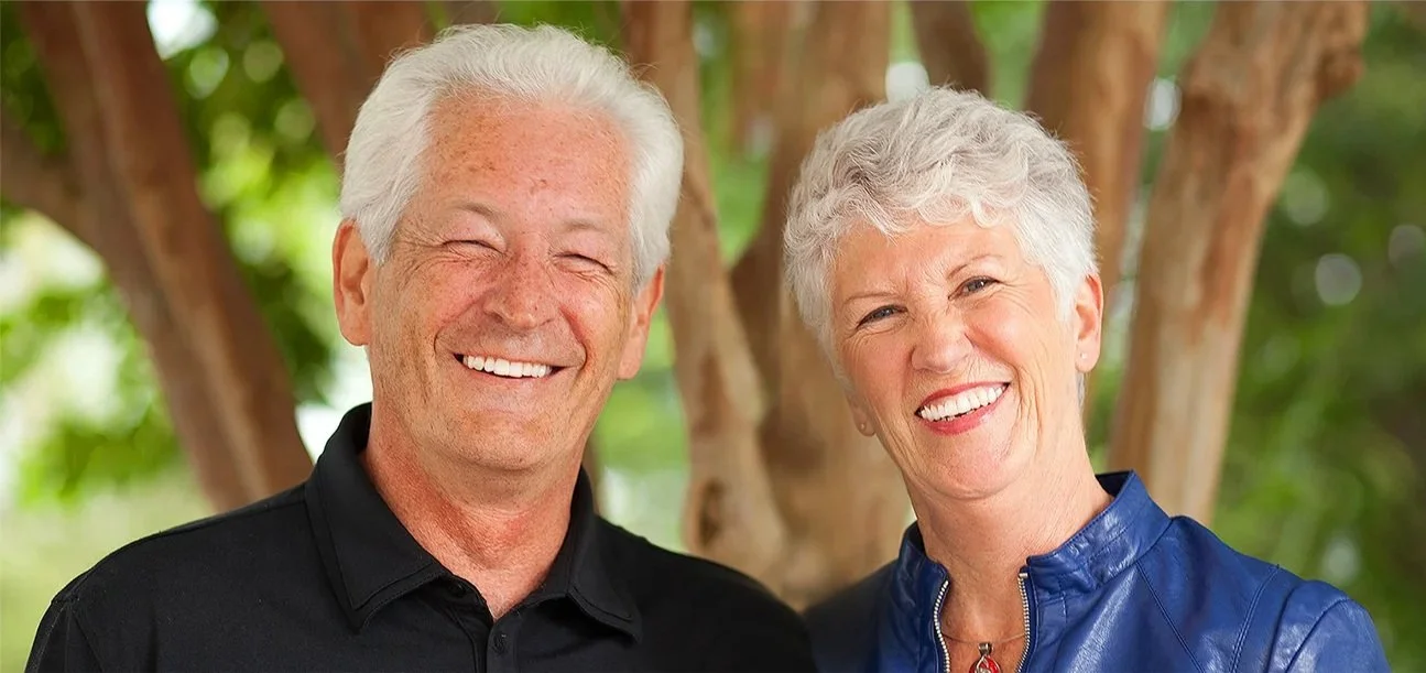 A smiling elderly couple standing outdoors in front of a tree with green foliage, enjoying a sunny day.