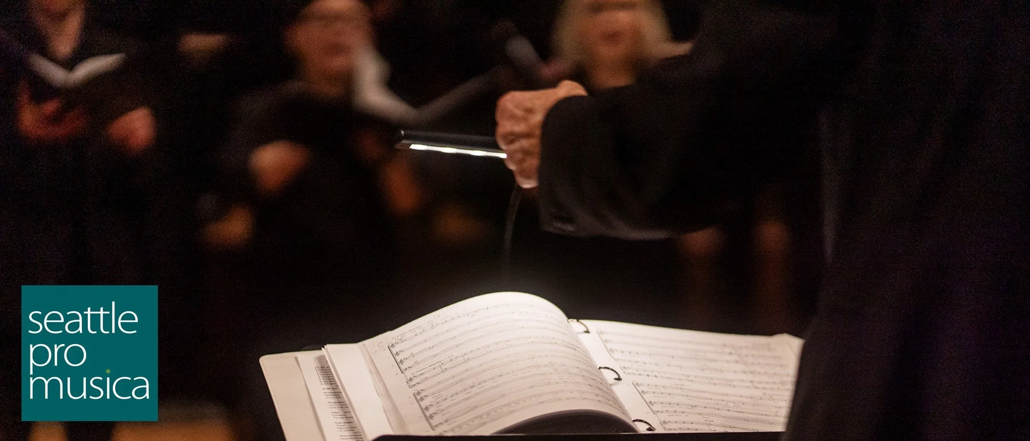 A choral concert. A musical score is lying on the conductor's podium. Choral singers are in the background. Logo with the words "seattle pro musica."