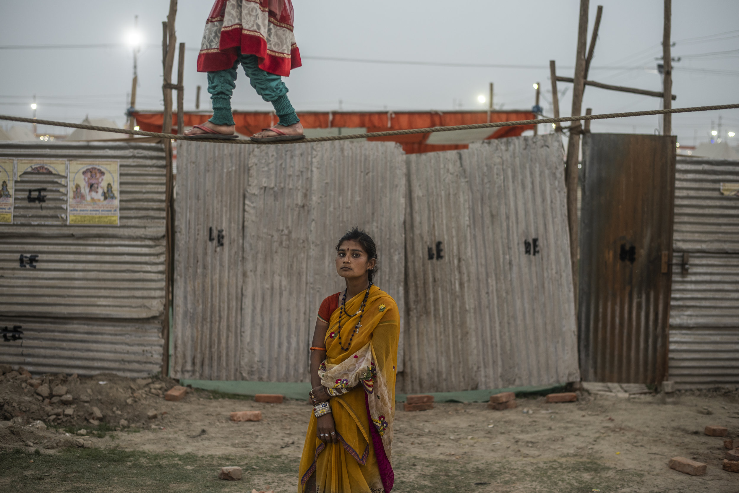  A young girl balances on a tight rope while another stands stoically below to collect money from bystanders. 