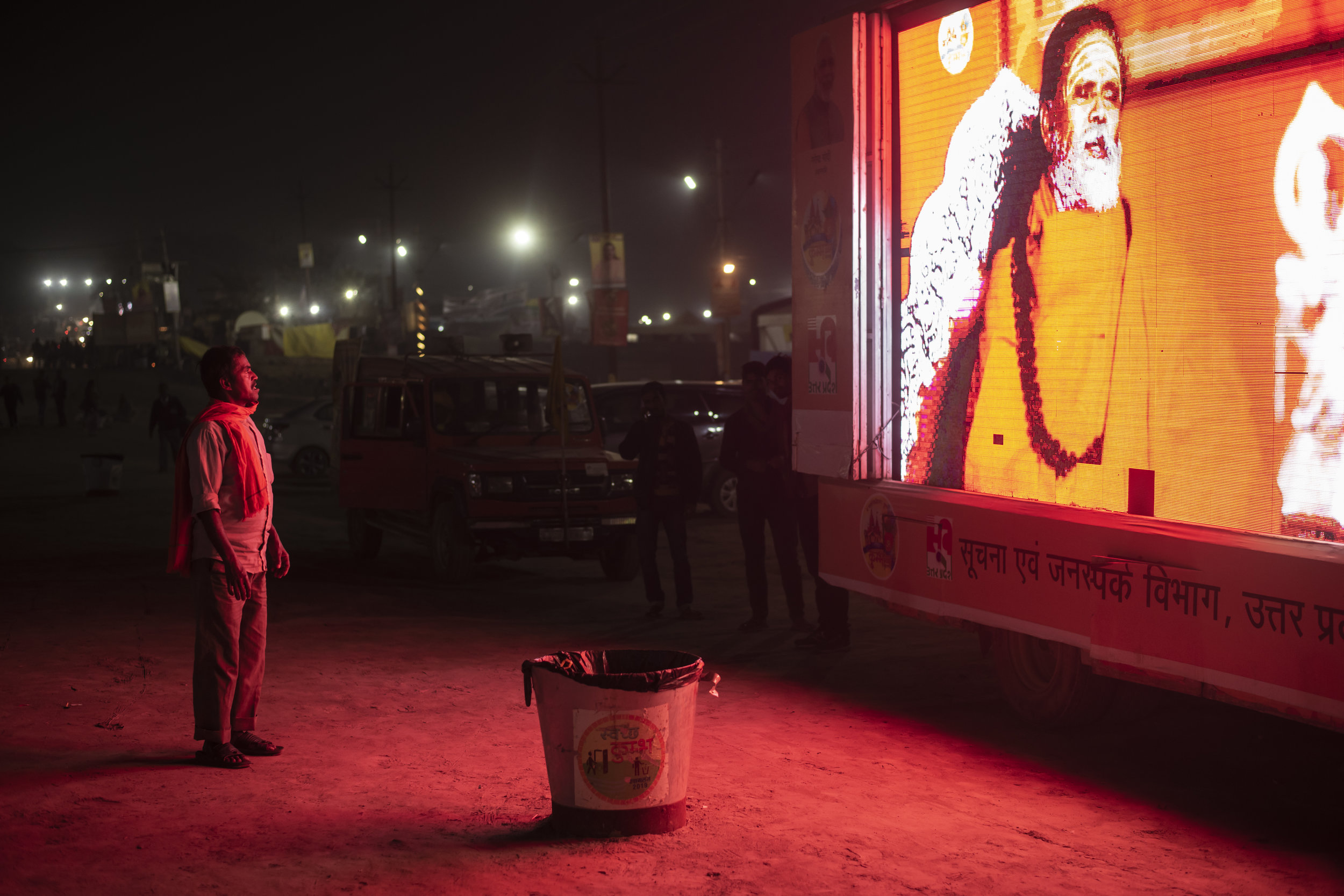  A man stands, captured in crimson light, downloading the teachings from the glowing roadside sadhu. Here at Kumbh consciousness moves. It is pulled and pushed. For some it shrinks and others it expands. 