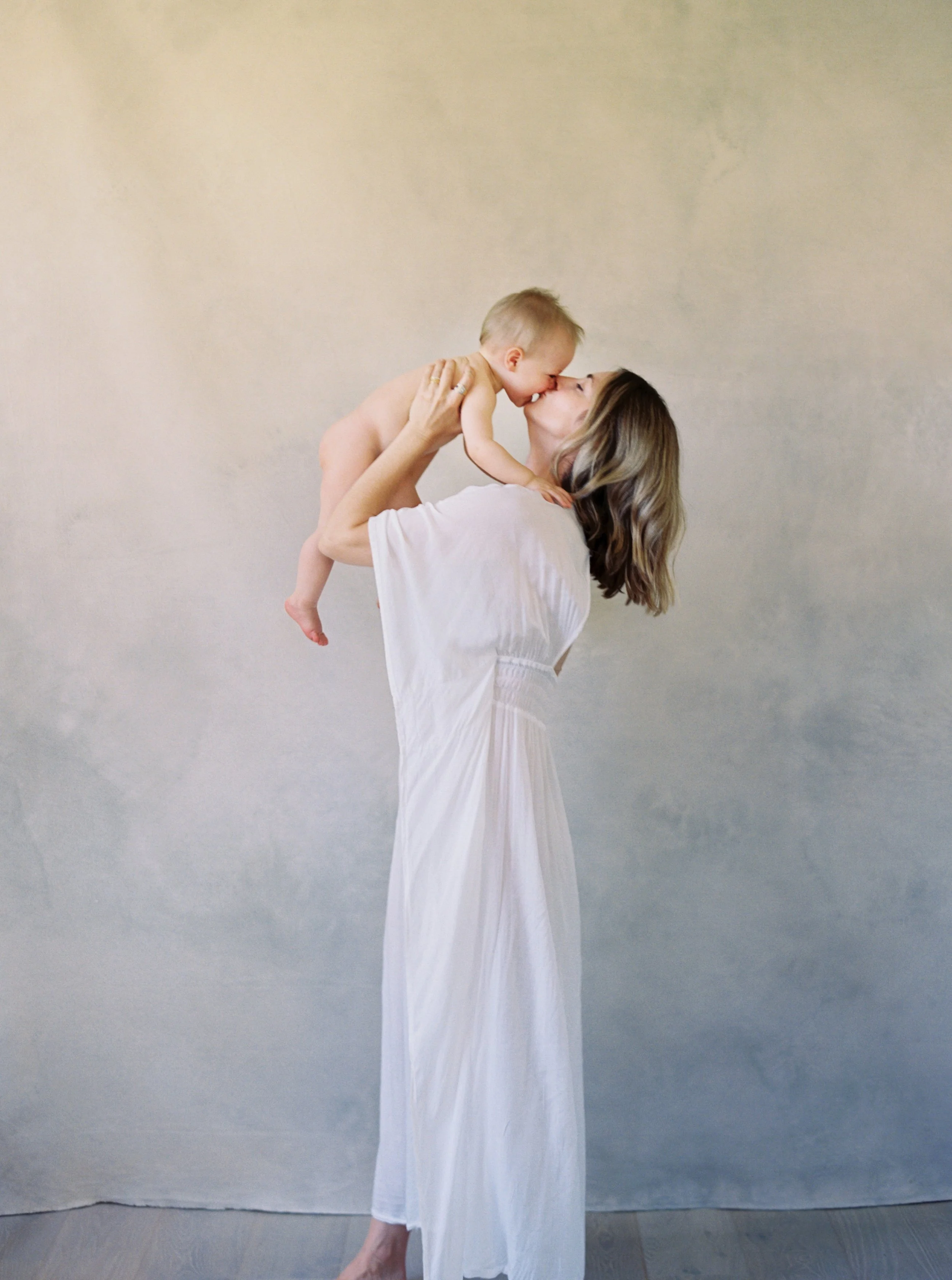 mother in white dress holding baby girl in the air and kissing her.