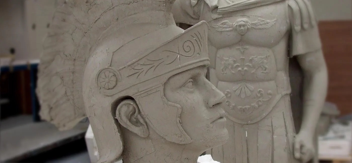 Close-up of a stone sculpture of a Roman soldier wearing a helmet with decorative details, with another partially visible sculpture in the background.