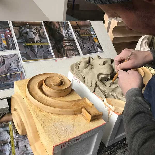 A person sculpting a lion's head in clay, with reference photos of a lion statue on the wall behind them, and a wooden amulet or decorative piece with a spiral design on the desk in front.