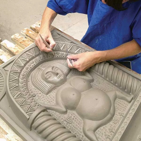 A person in a blue shirt is carving a detailed sculpture of a reclining woman with large breasts from a gray stone or clay tray.