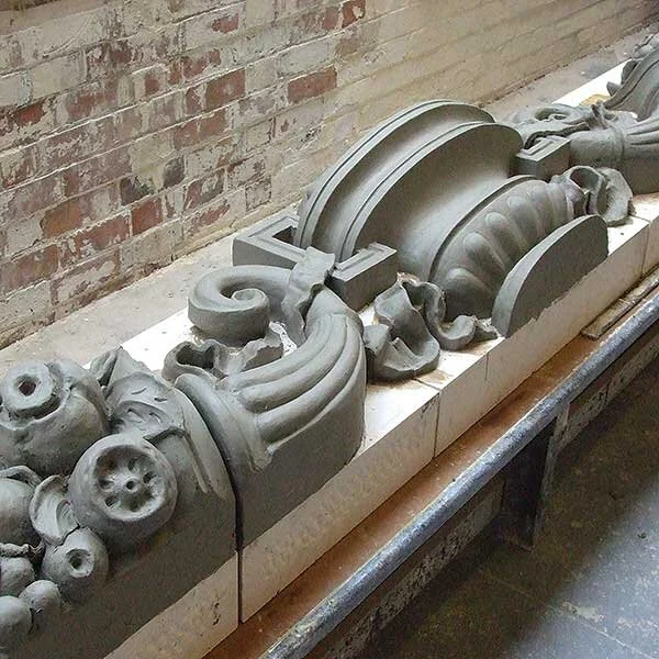 Close-up of decorative architectural stone carvings, including a shell motif and curved ornamental designs, placed on a workbench against a brick wall.